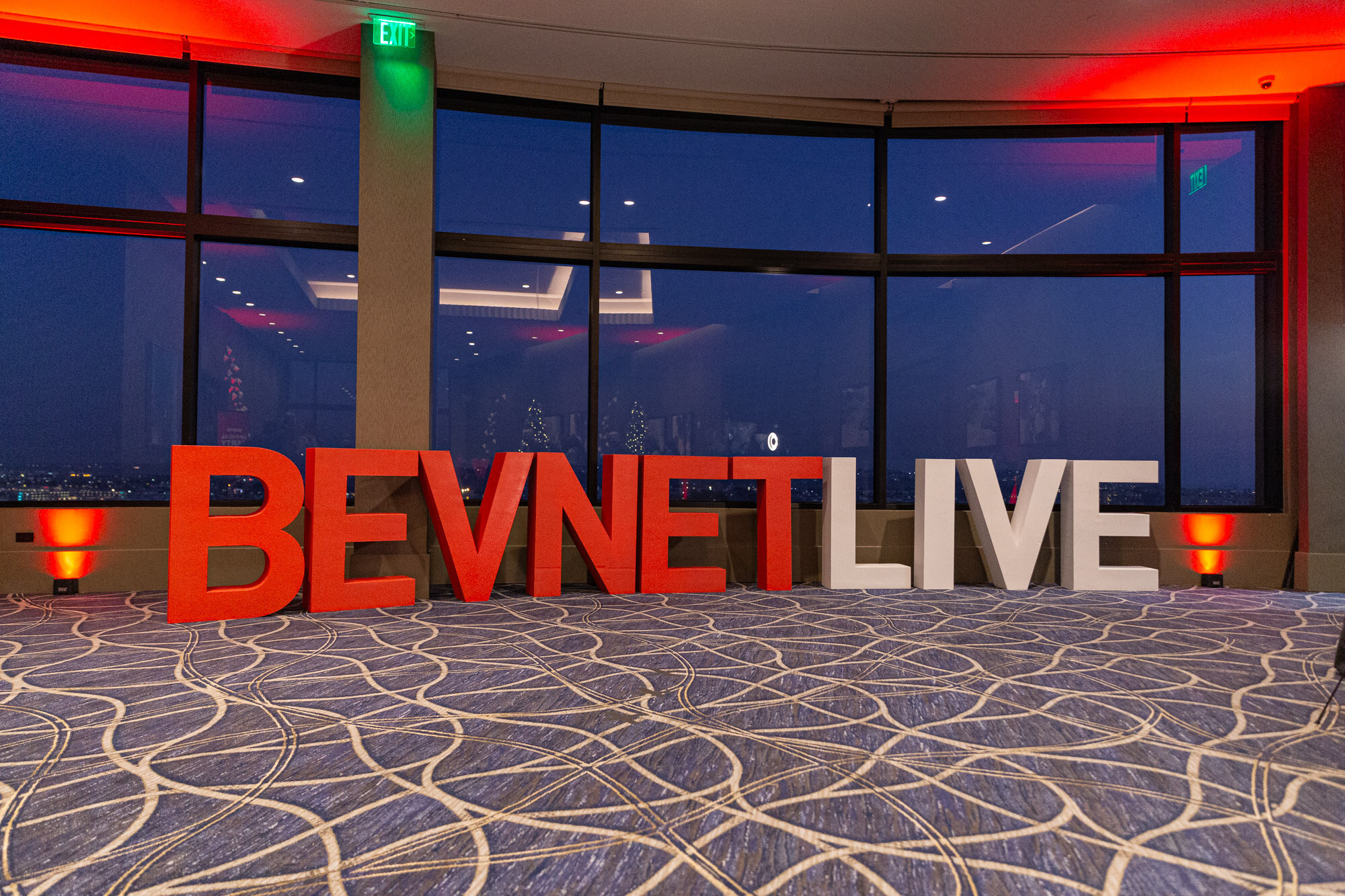 Large red and white BEVNET LIVE letters on a patterned carpet in front of dark windows with red lighting.