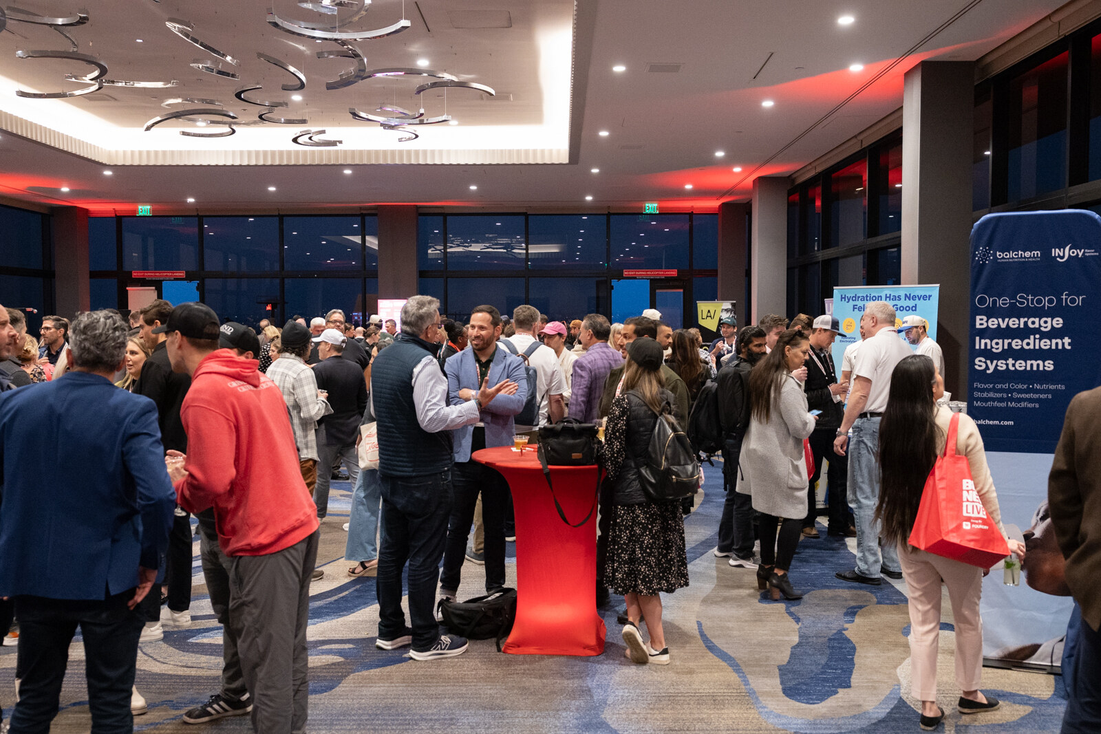 A crowd stands in a large rooftop ballroom, some holding red bags, near sample tables.
