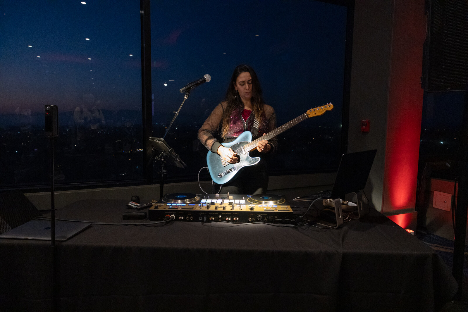 Woman plays a light blue electric guitar behind a DJ table with a microphone in front of a dark window.