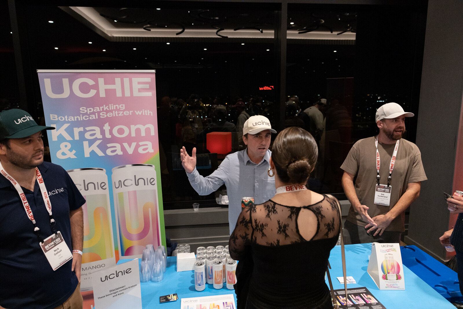 People stand behind a table with an Uchie banner and cans in front of a window at night.