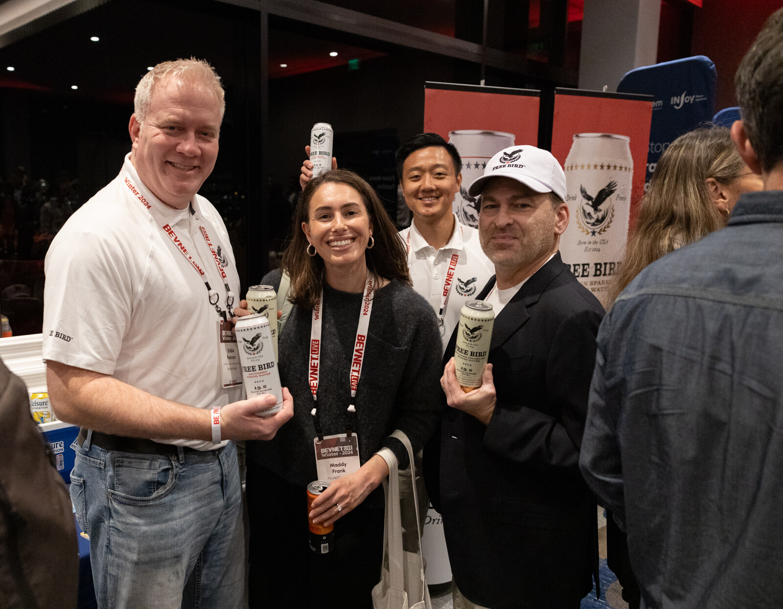 Four people, one in a white cap holding a can, smile at the camera at a sampling table with red banners behind them.
