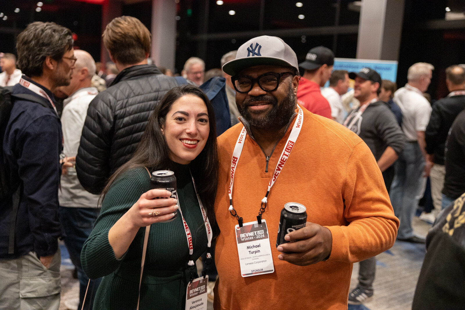 A woman in green and a man in an orange sweater hold cans and smile in a crowded room.