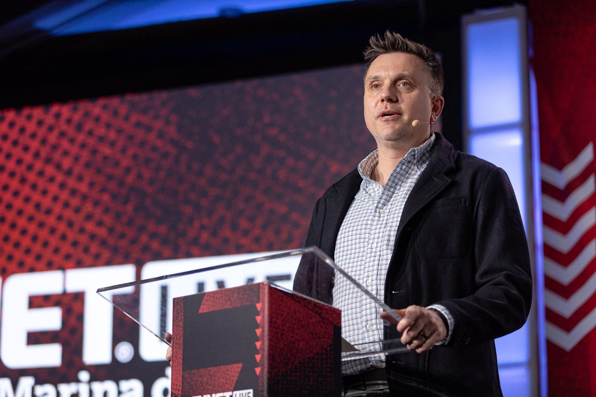 Man in a dark jacket and checked shirt speaks at a podium in front of a BevNET Live Marina del Rey screen.