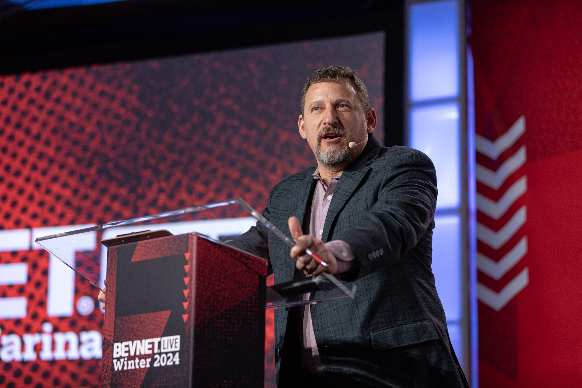 Bearded man in a dark blazer gestures at a podium that reads BevNET Live Winter 2024.