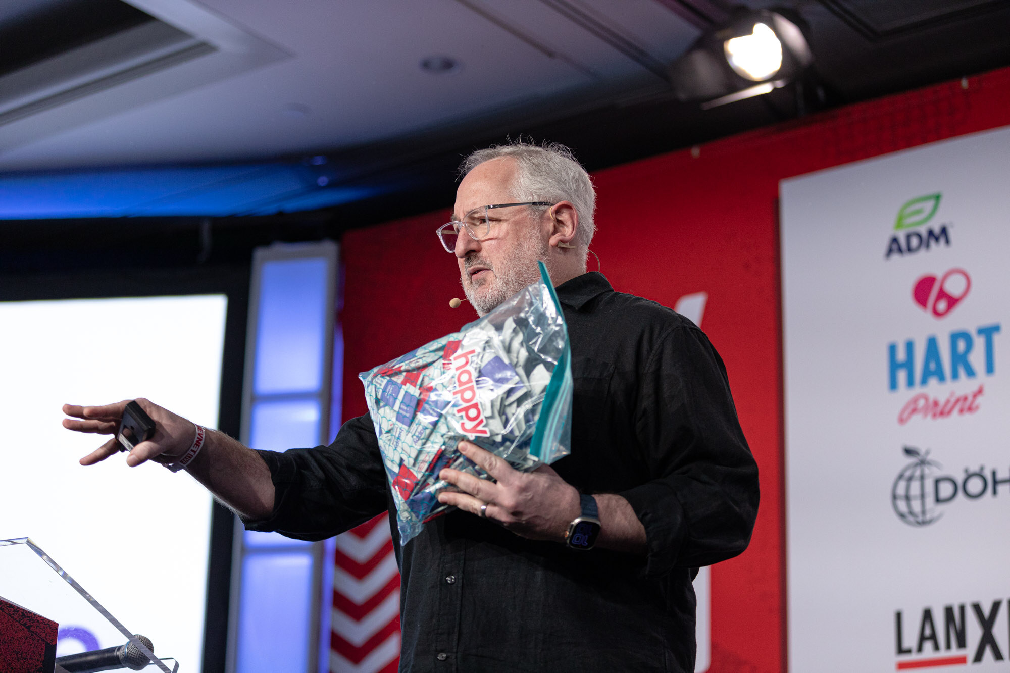 Man with glasses and a black shirt holds a clear bag of colorful items in one hand and gestures with the other.