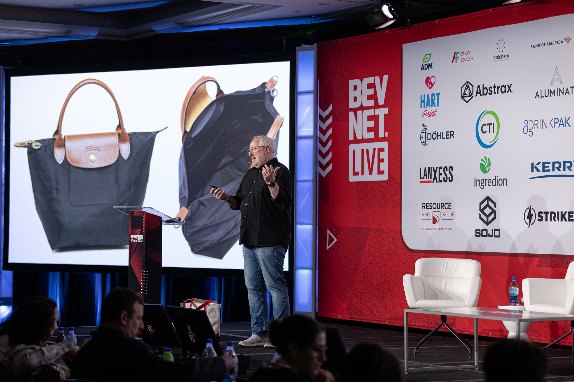Man in a black shirt stands on stage beside a screen showing a navy handbag with a tan handle.