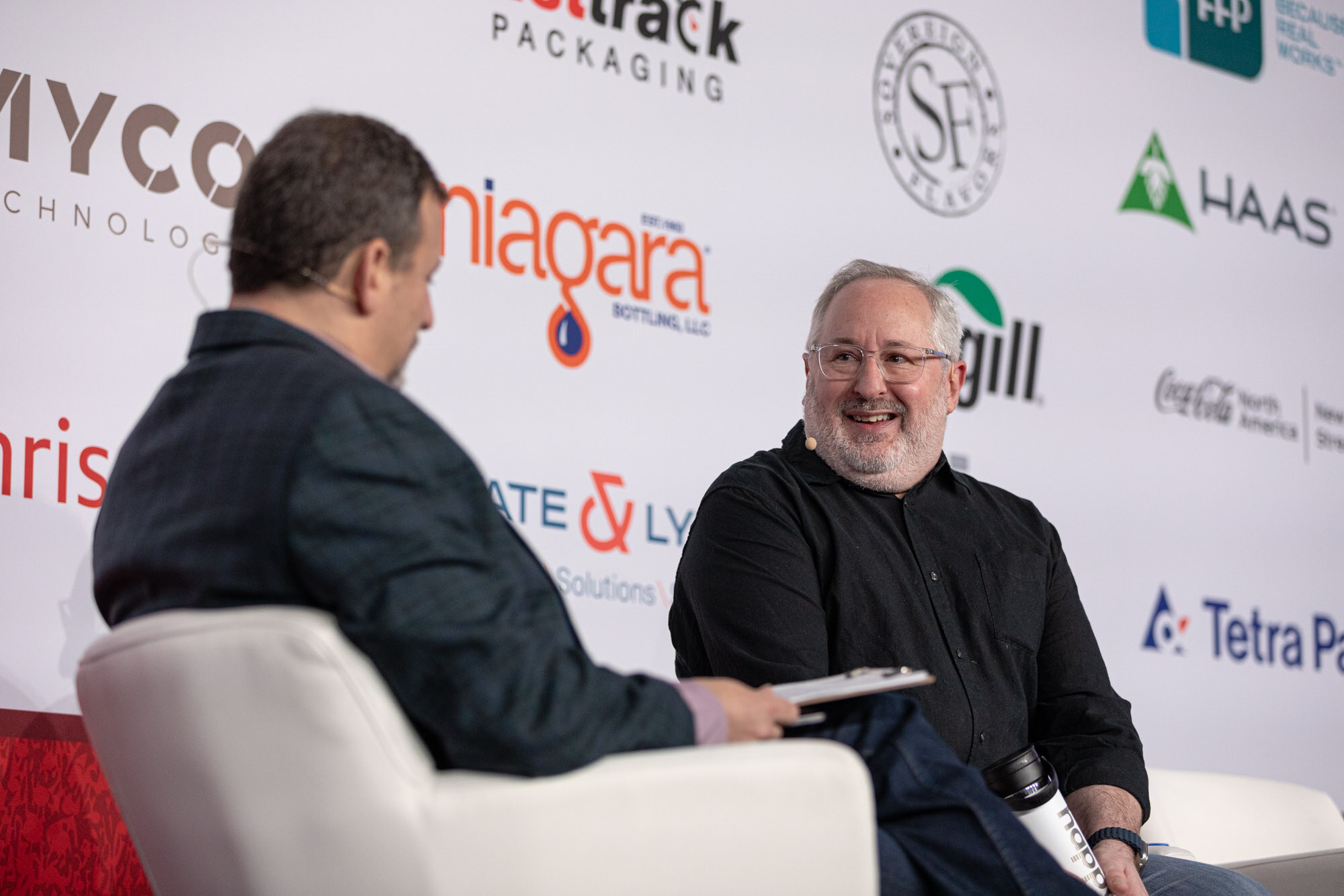 A man in a dark jacket and a smiling man in black sit in white armchairs on stage.