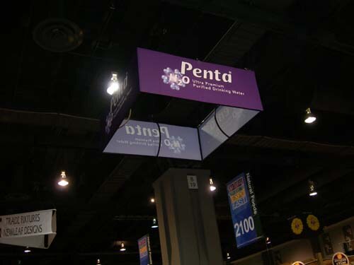 Purple hanging sign reading Penta Ultra Premium Purified Drinking Water above a column in an exhibit hall, with a Natural Foods 2100 banner nearby.