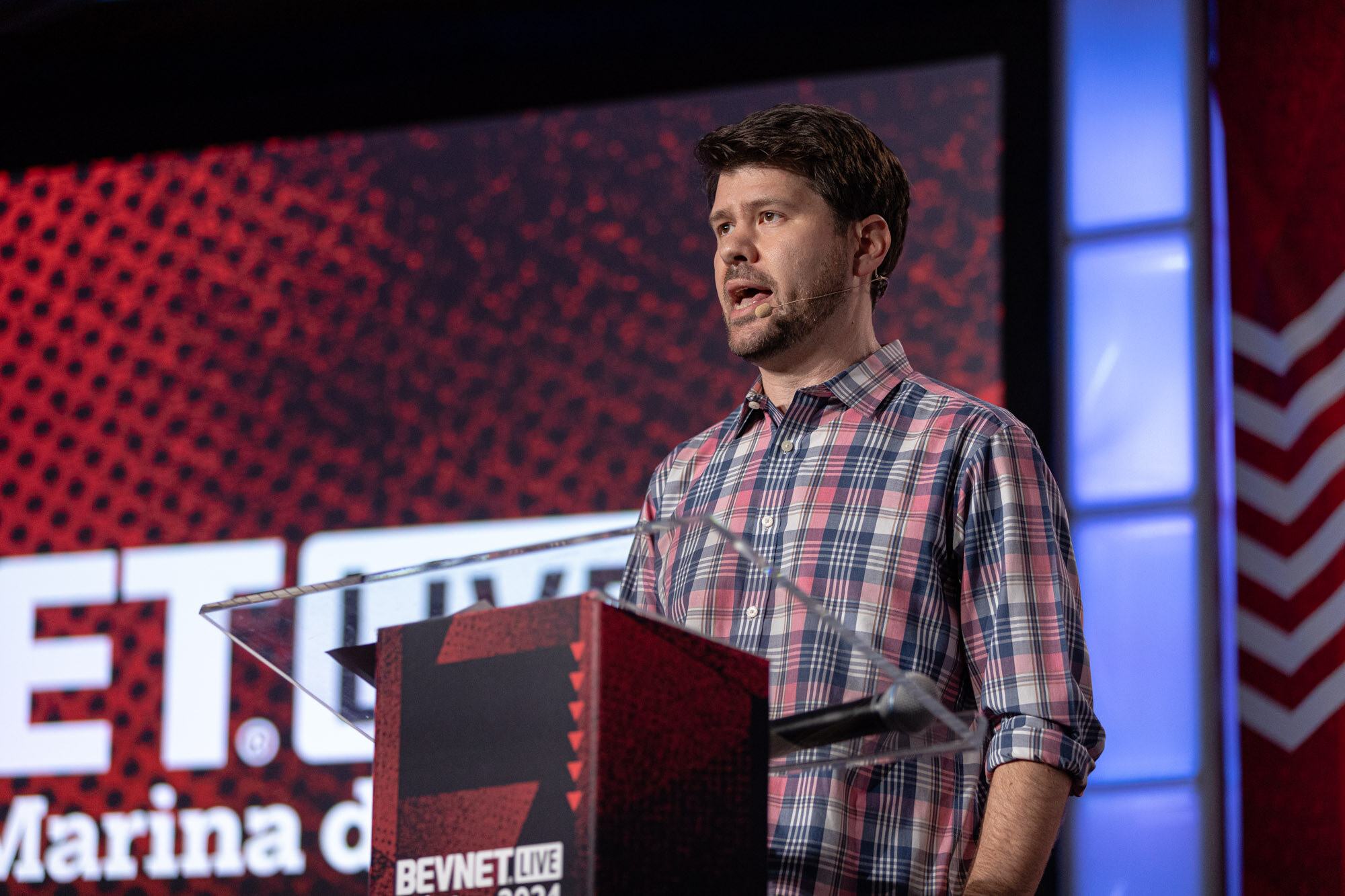 Man in a plaid shirt speaks at a podium in front of a red BevNET Live backdrop.