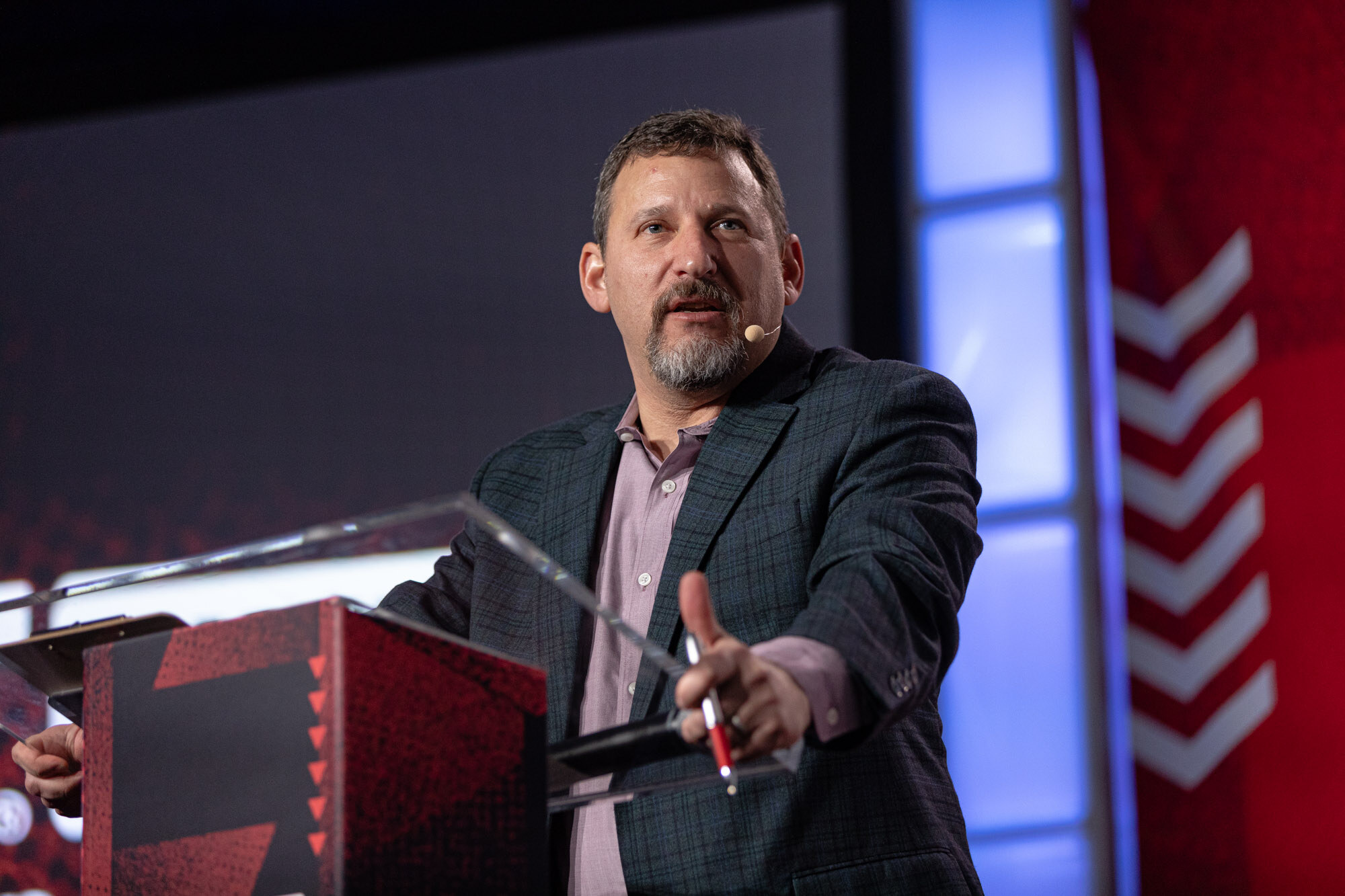 Bearded man in a dark blazer gestures at a podium in front of a red stage backdrop.