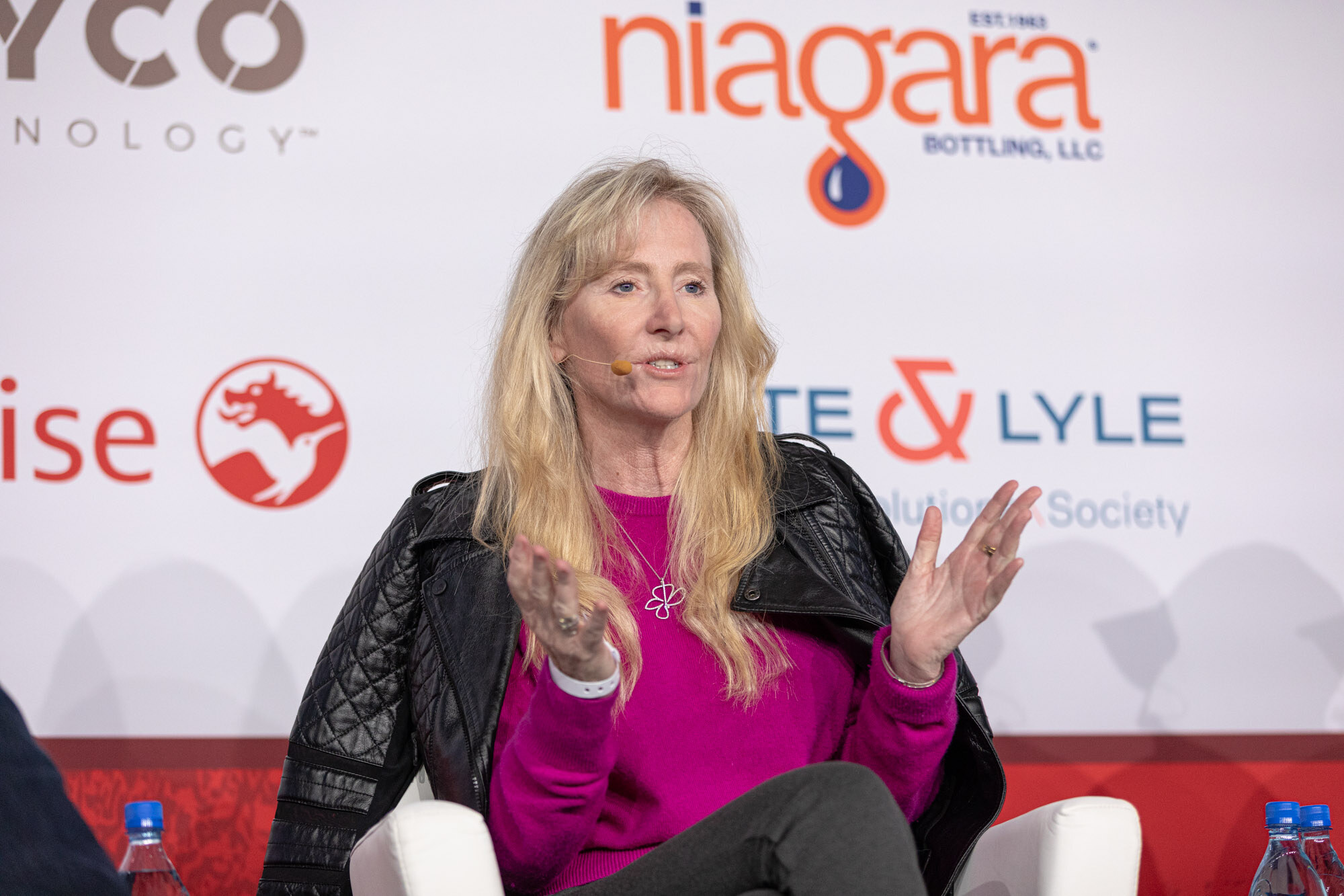 Woman with long blond hair in a magenta top and black jacket gestures while seated in front of a sponsor wall.