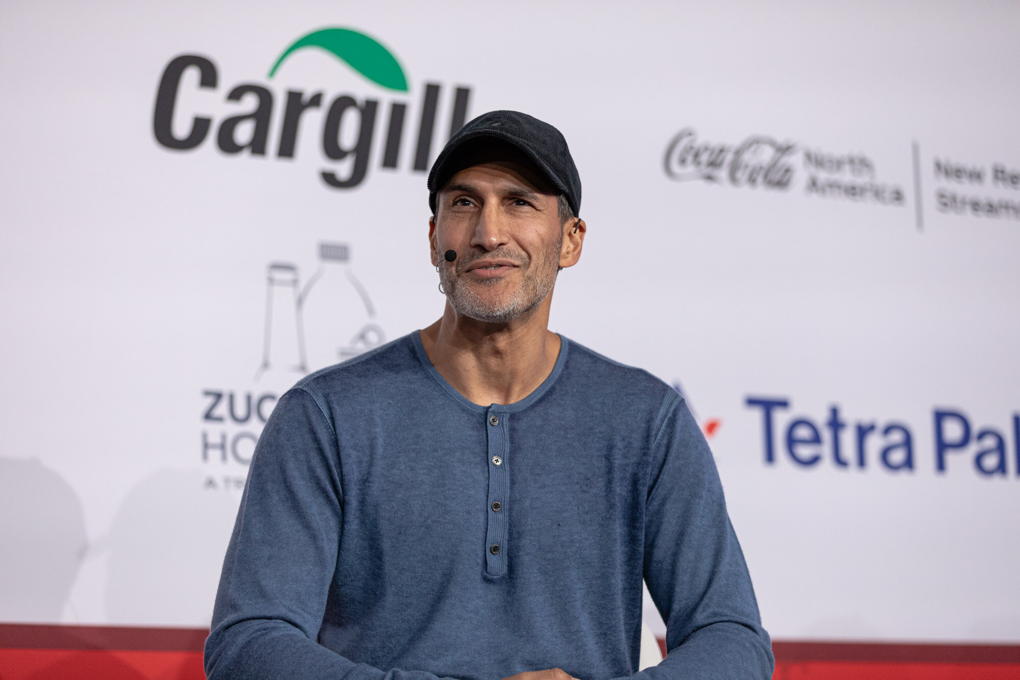 Man in a black cap and blue henley shirt sits in front of a Cargill and Coca-Cola North America sponsor wall.