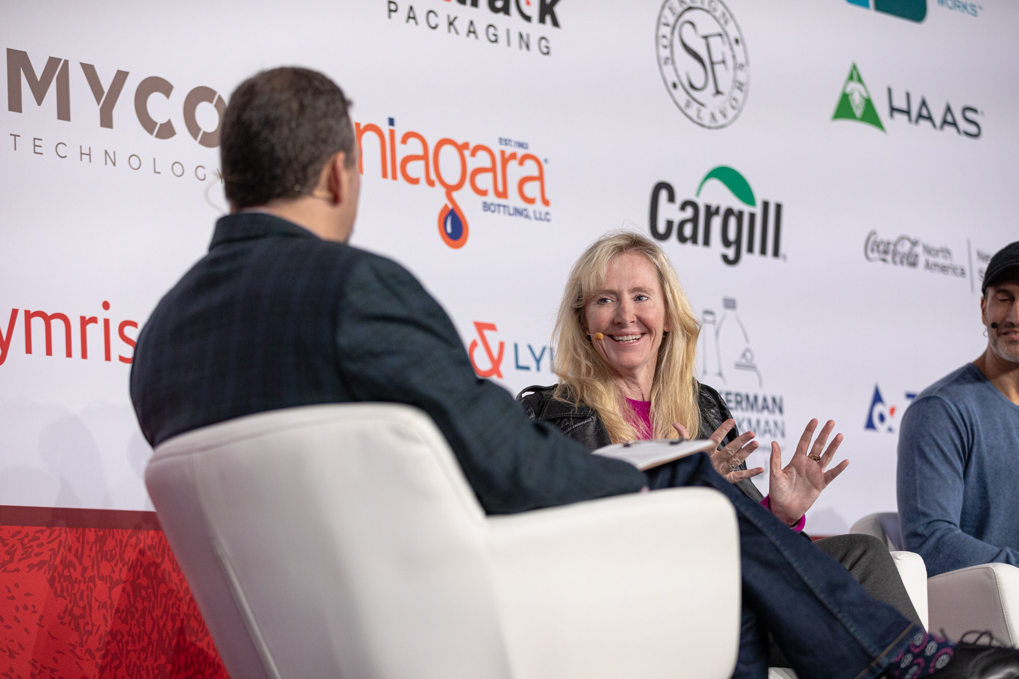 A blond woman gestures from a white armchair while the moderator sits with his back to the camera.
