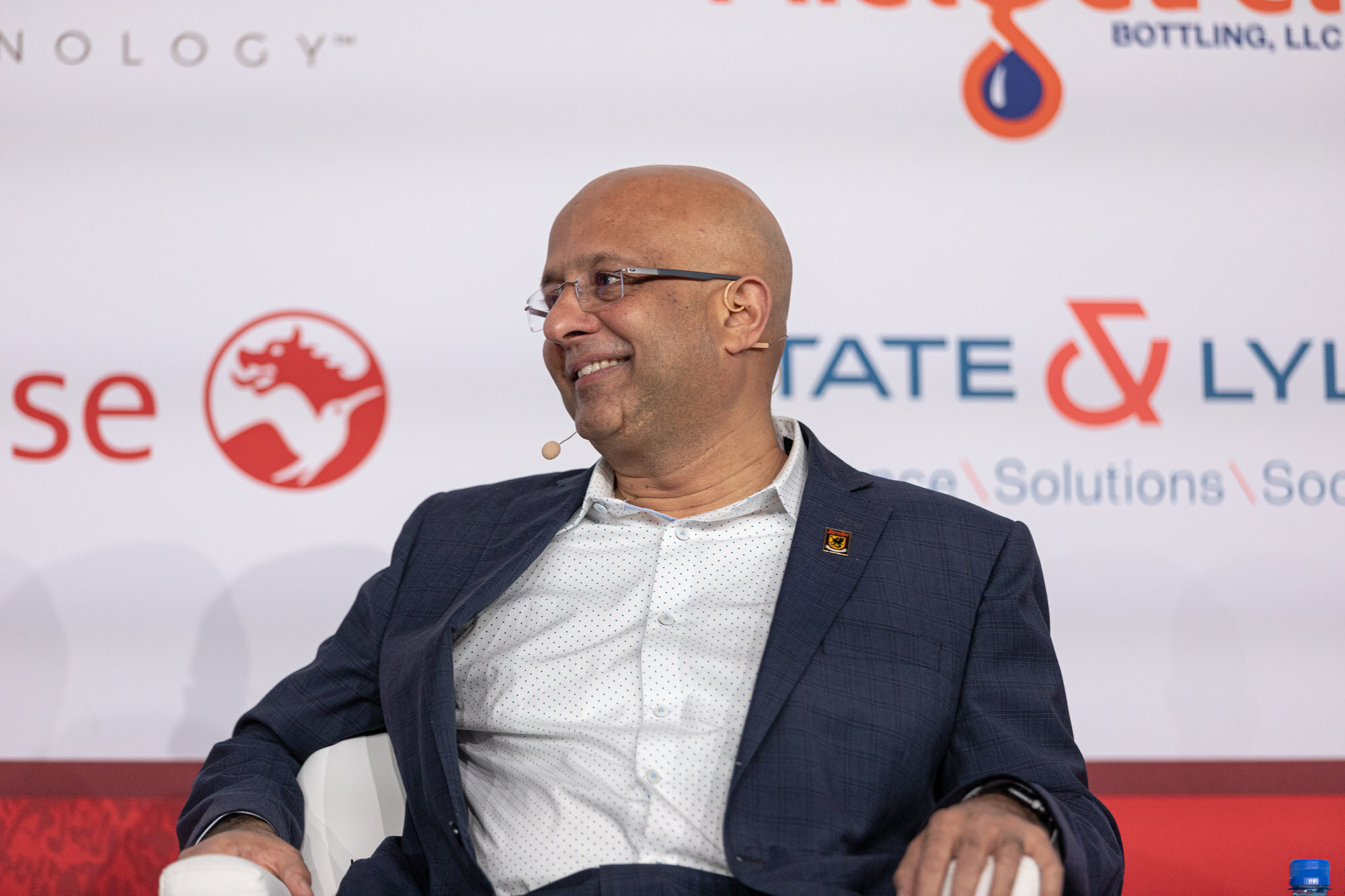 Bald man with glasses in a navy blazer smiles broadly while seated in front of a sponsor wall.