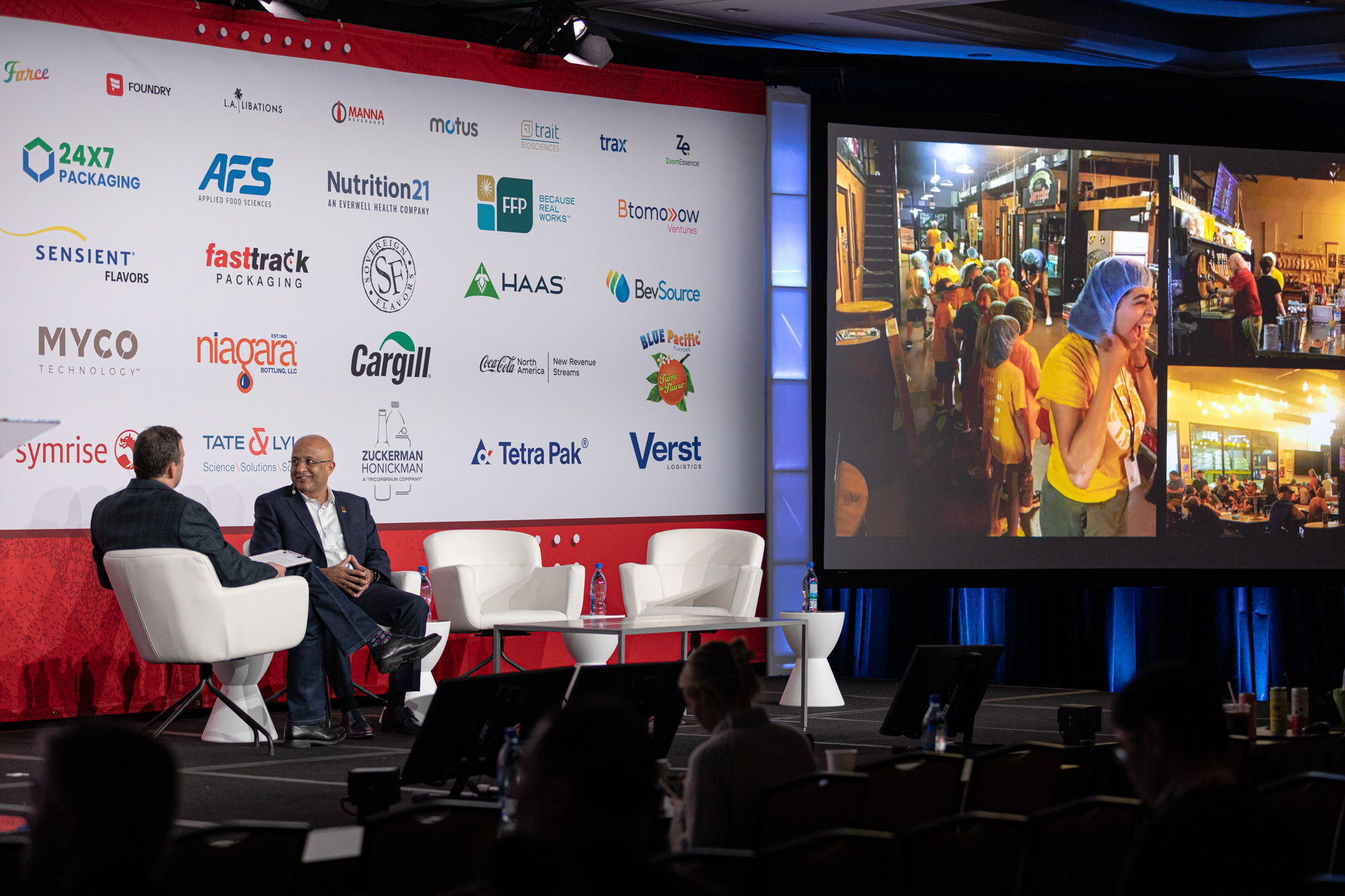 Two men sit in white chairs on stage next to a big screen showing a street scene.