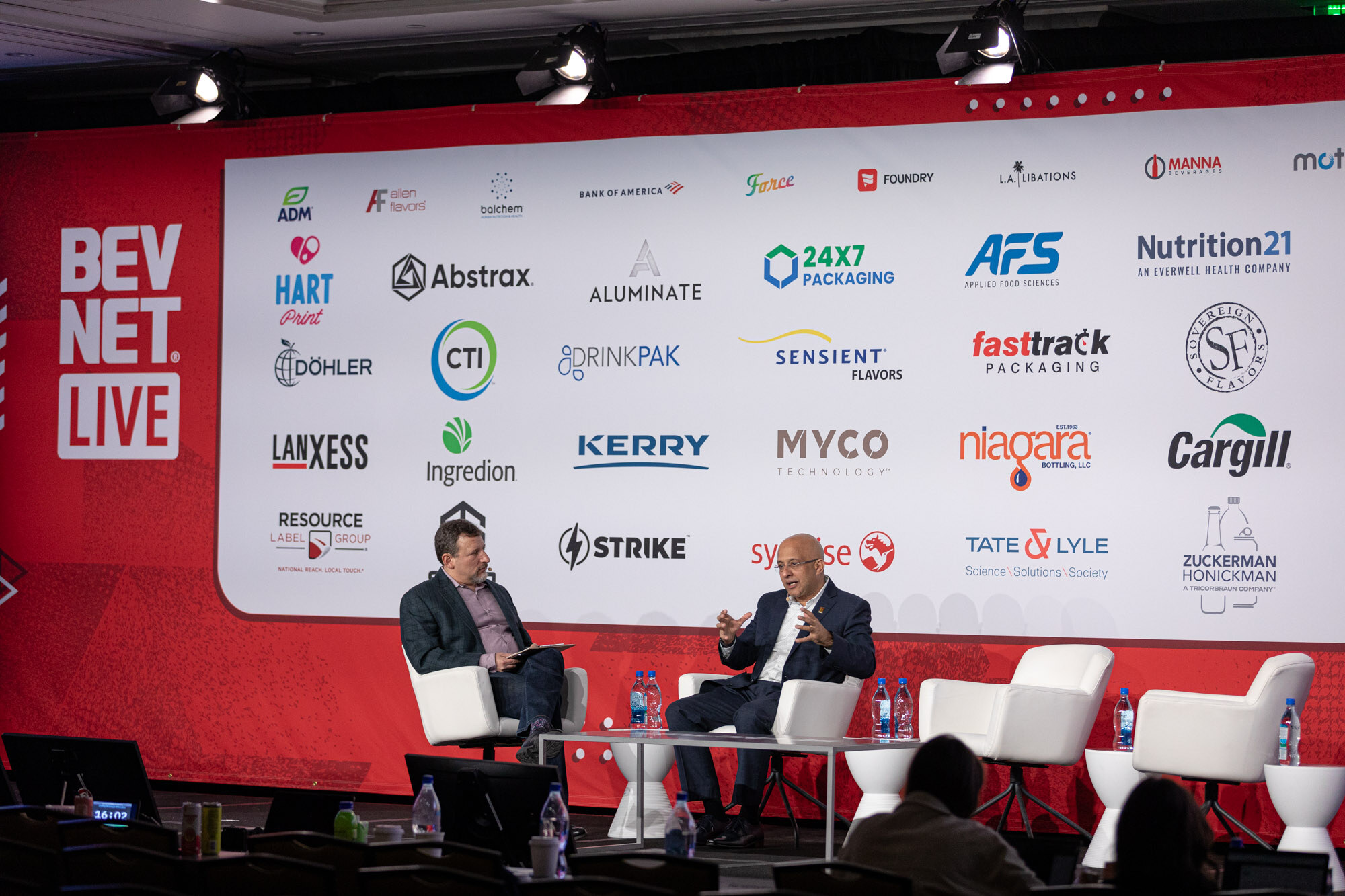 Two men sit in white chairs on a stage in front of a large sponsor wall.