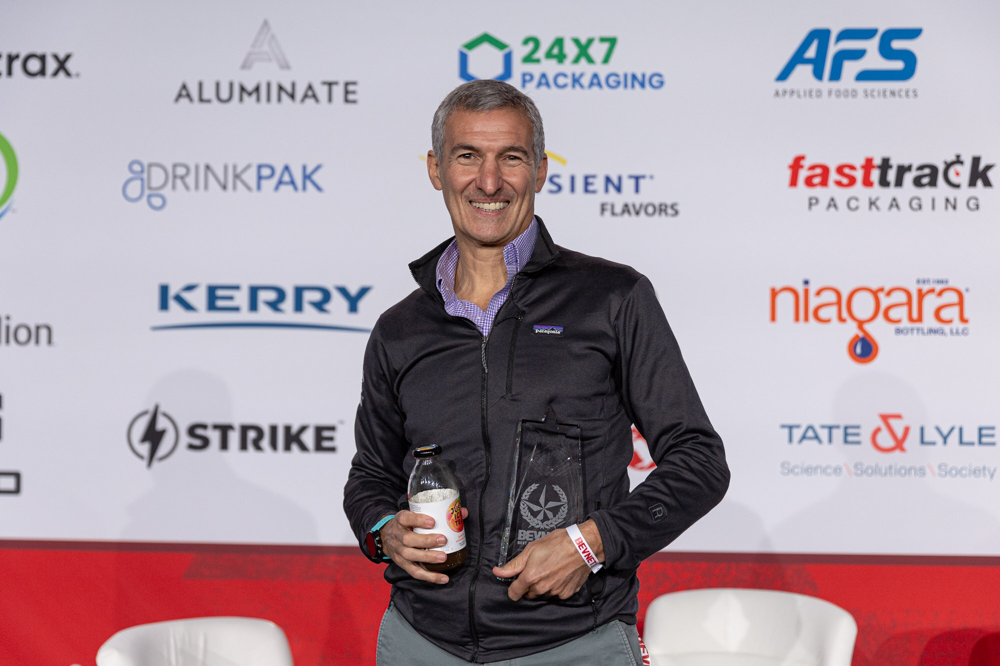 Smiling man in a black jacket holds a glass trophy and a small bottle in front of a sponsor wall.