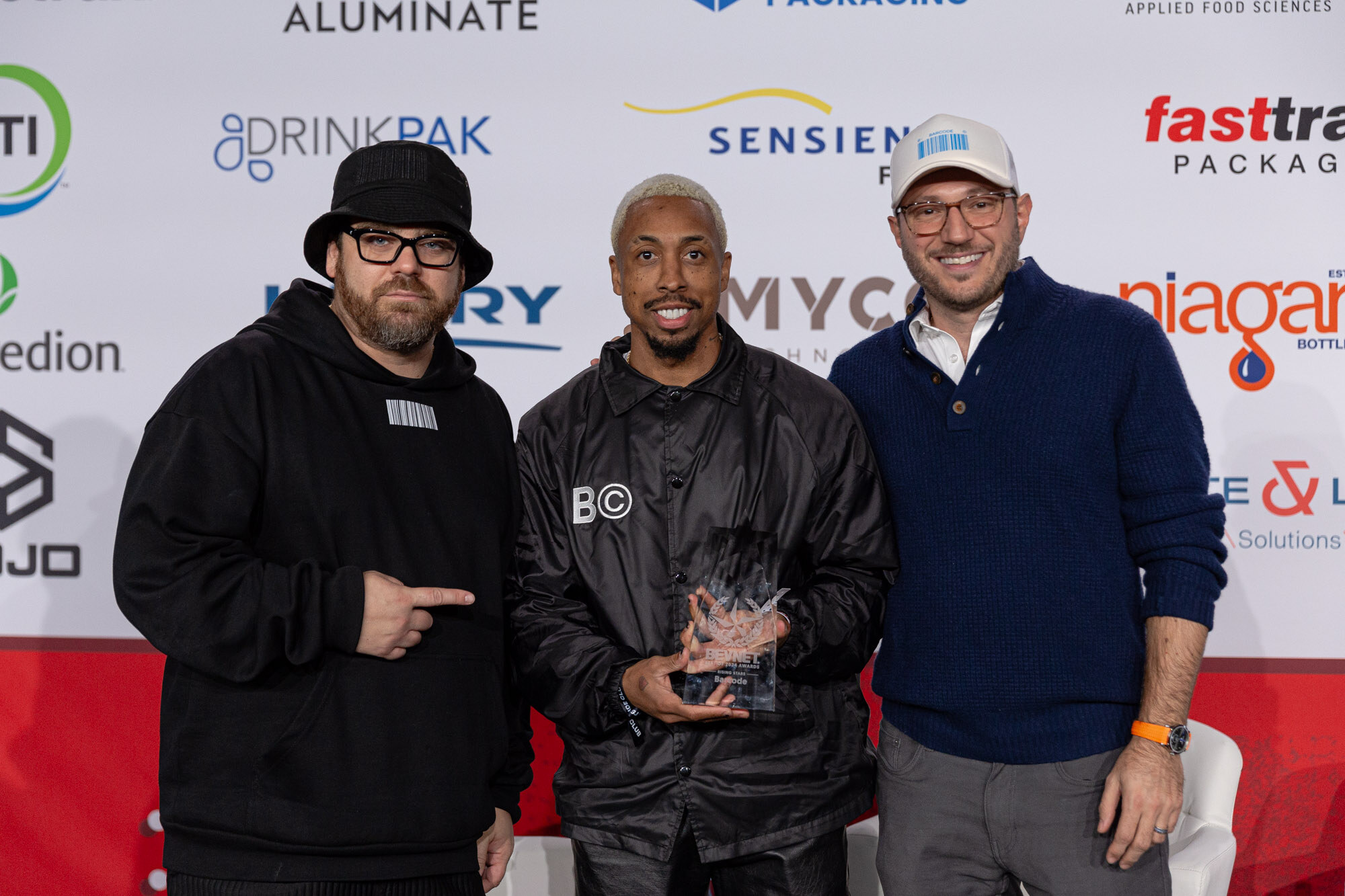 Three men stand together, one in a black hoodie pointing at the man in the middle who holds a glass trophy.