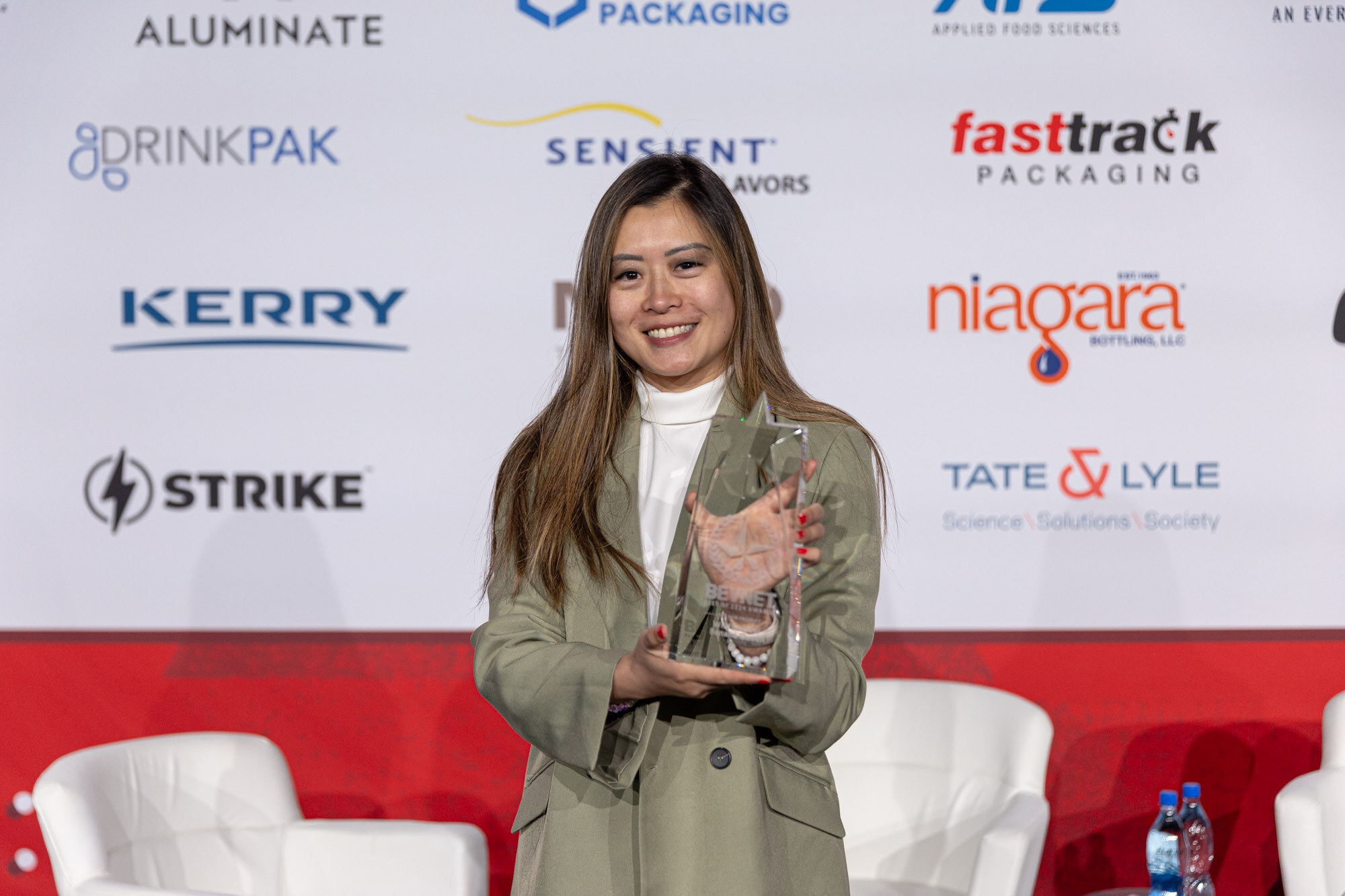 Woman in an olive blazer and white turtleneck holds a glass trophy in front of a sponsor wall.