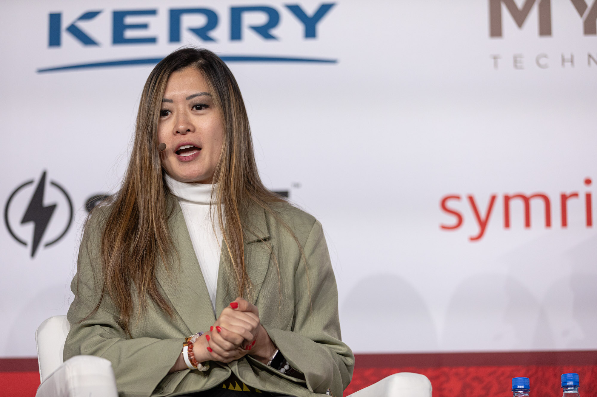 Woman with long brown hair in a green blazer and white top speaks while seated in front of a Kerry sponsor banner.
