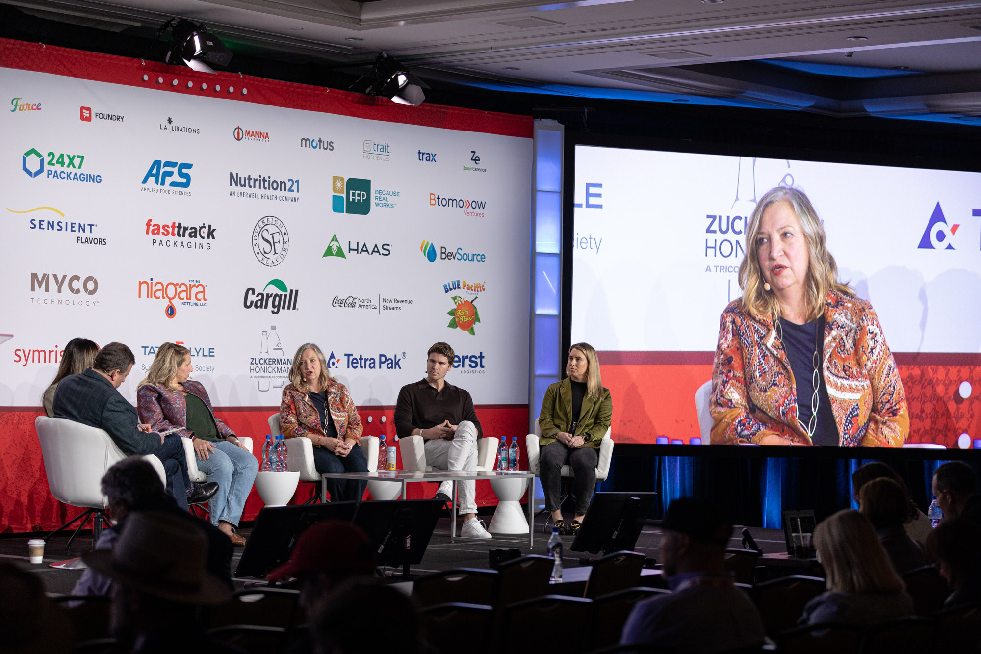 Six people sit in a row on stage in front of a sponsor wall and a large screen showing a close-up of a panelist.