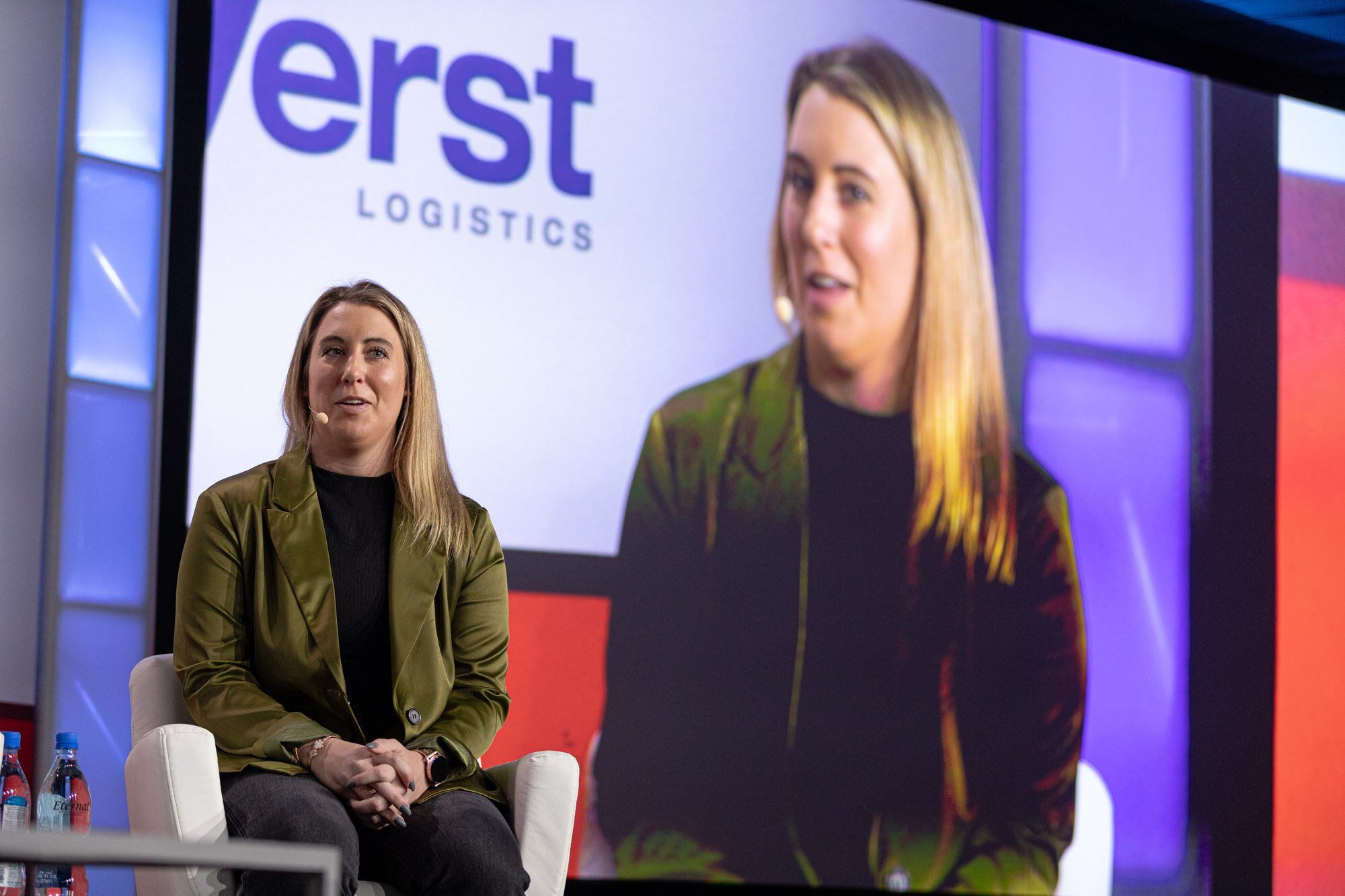 Woman in an olive blazer sits on stage in front of a large screen showing her close-up with the Verst Logistics logo.