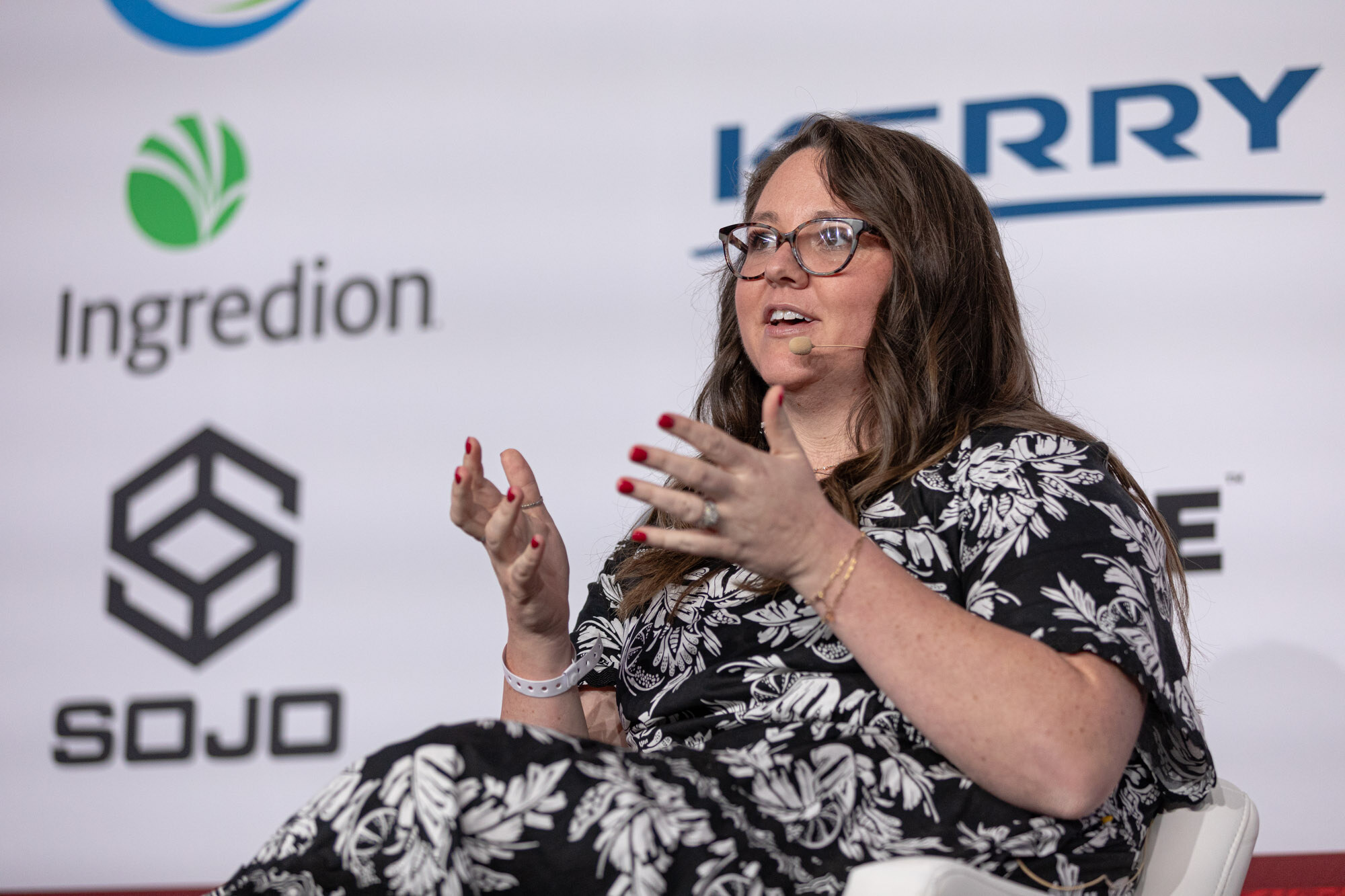 Woman with dark-framed glasses in a black-and-white floral dress gestures in front of a Sojo sponsor banner.