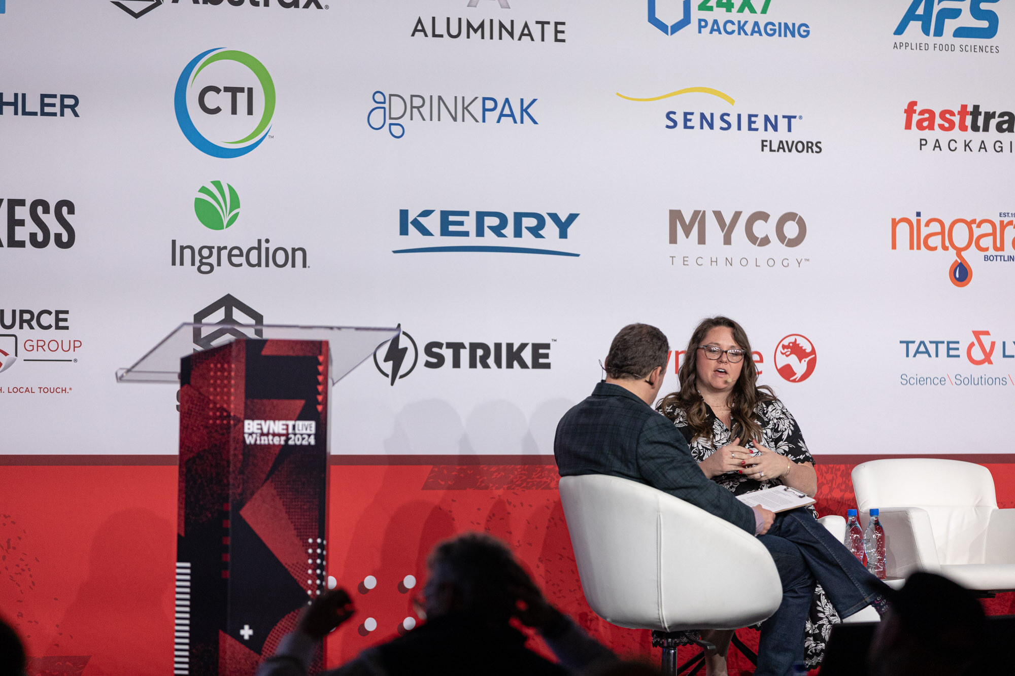 Two people sit in white armchairs at the right of a podium that reads BevNET Live.