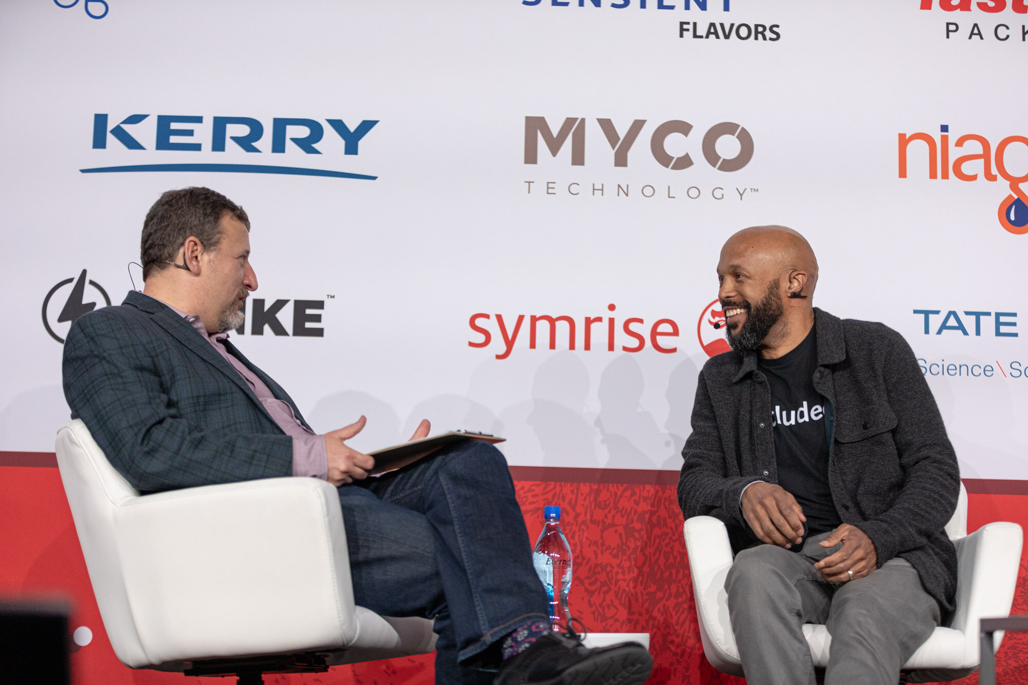 A man in a dark jacket and a laughing man in a black jacket sit in white armchairs in front of a sponsor wall.