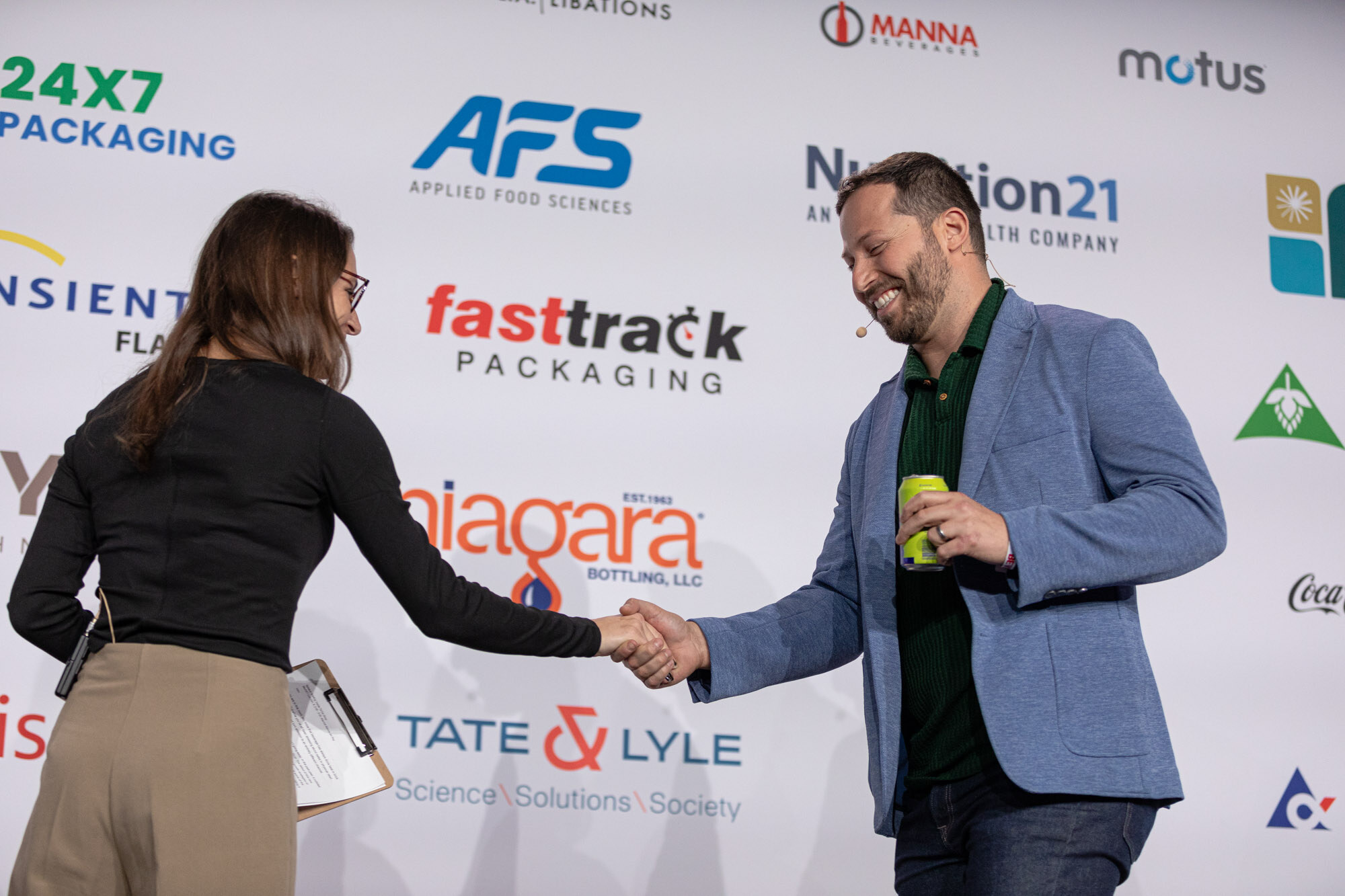 A woman in black and a man in a blue blazer shake hands on stage in front of a sponsor backdrop.