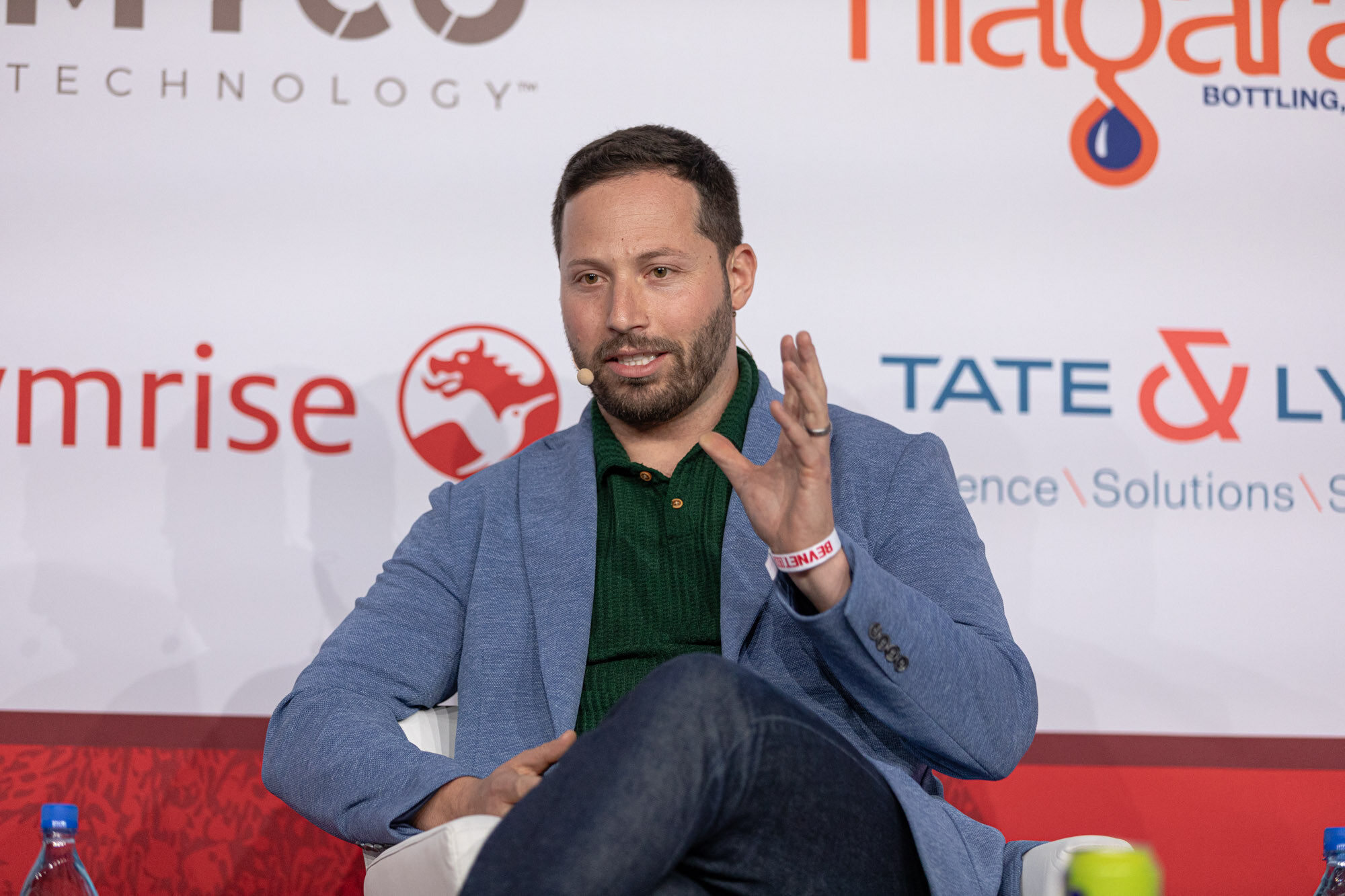 Man in a blue blazer and green shirt gestures while seated on stage in front of a sponsor wall.