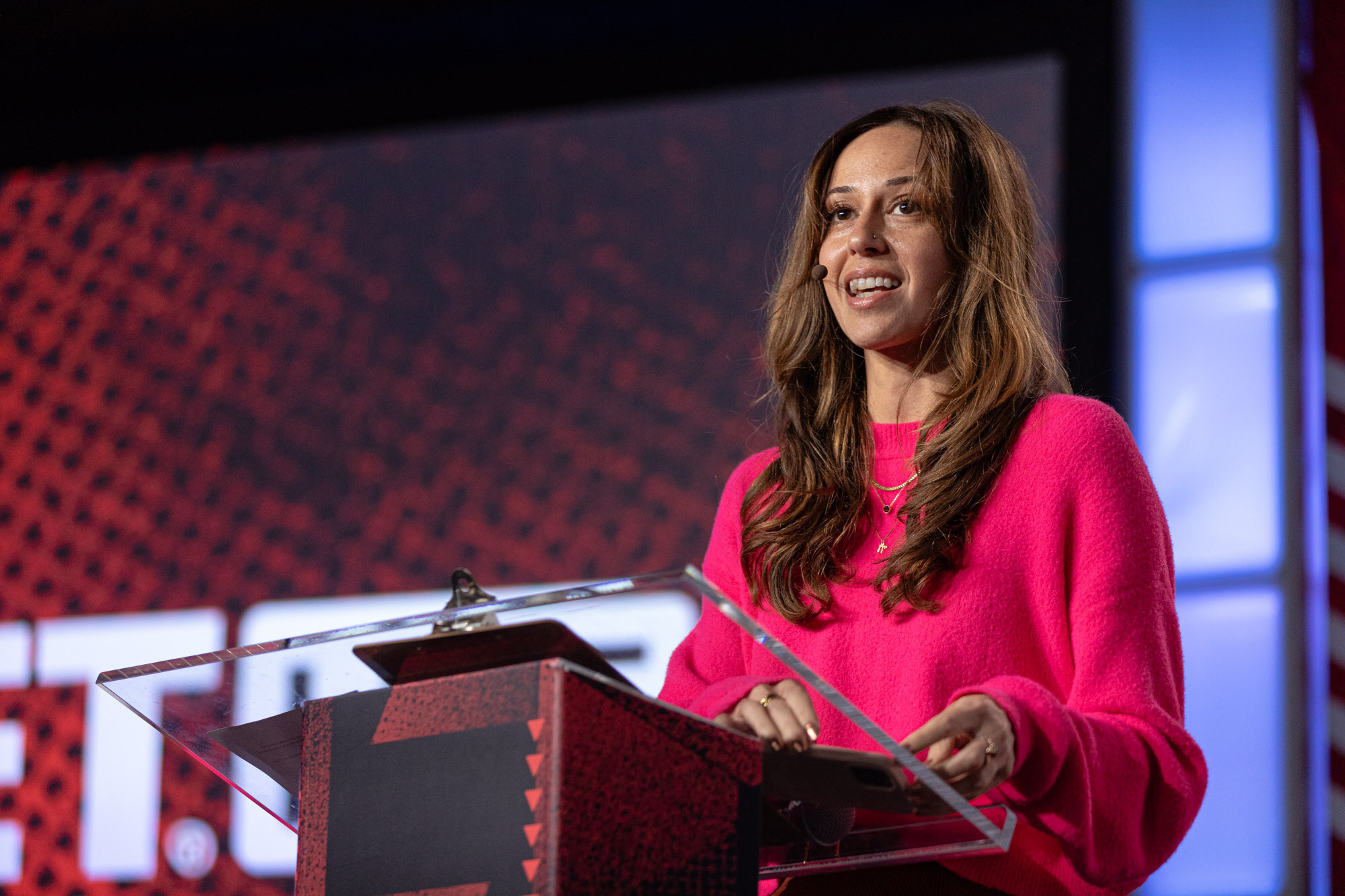 Smiling woman in a bright pink sweater speaks at a clear lectern against a dark red stage background.