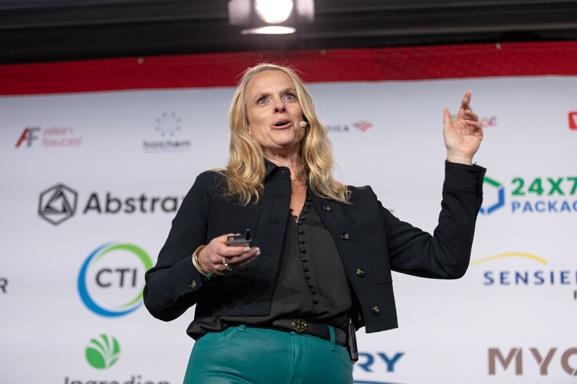 Woman with blond hair in a black jacket and teal pants gestures on stage in front of a sponsor wall.