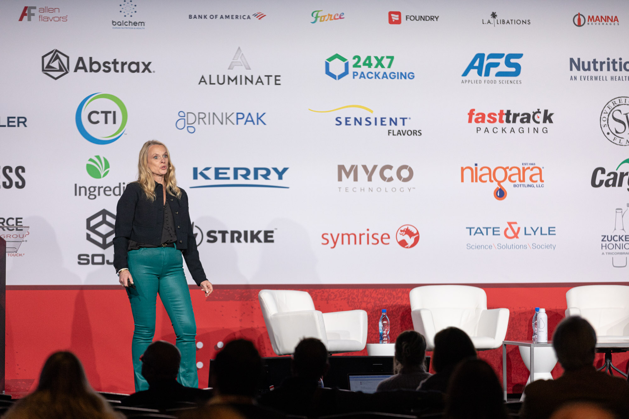 Speaker in teal pants stands on a stage in front of the sponsor backdrop with white chairs and the audience in the foreground.