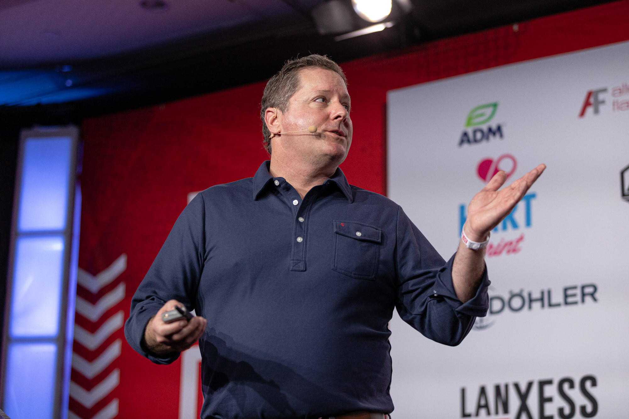 Man in a navy polo gestures on stage in front of a red BevNET Live backdrop.
