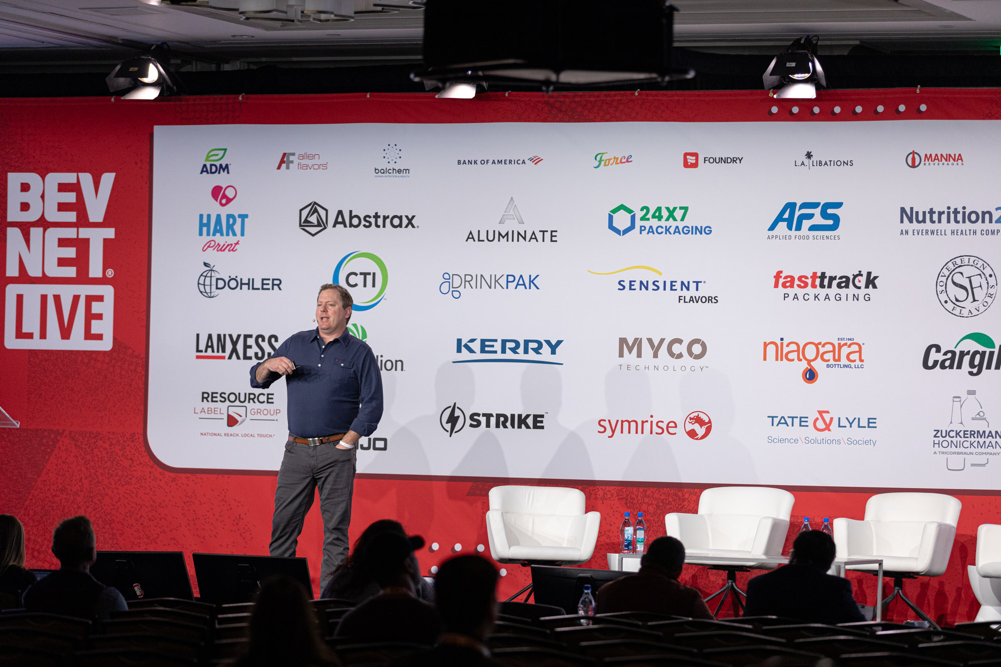 A man stands center stage in front of a large sponsor wall with white chairs and audience heads in silhouette.