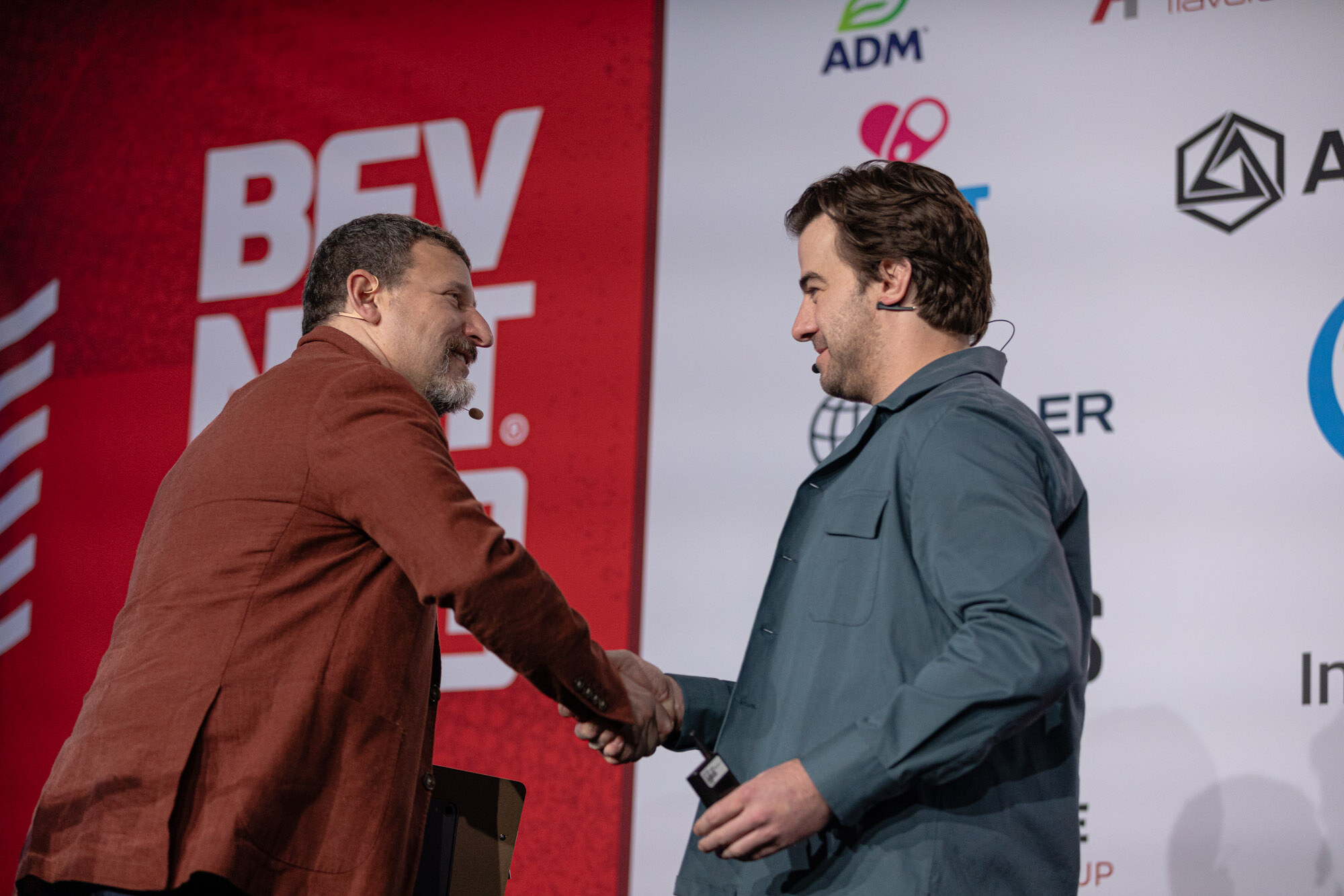 Two men in jackets shake hands on stage in front of a red BevNET Live backdrop.