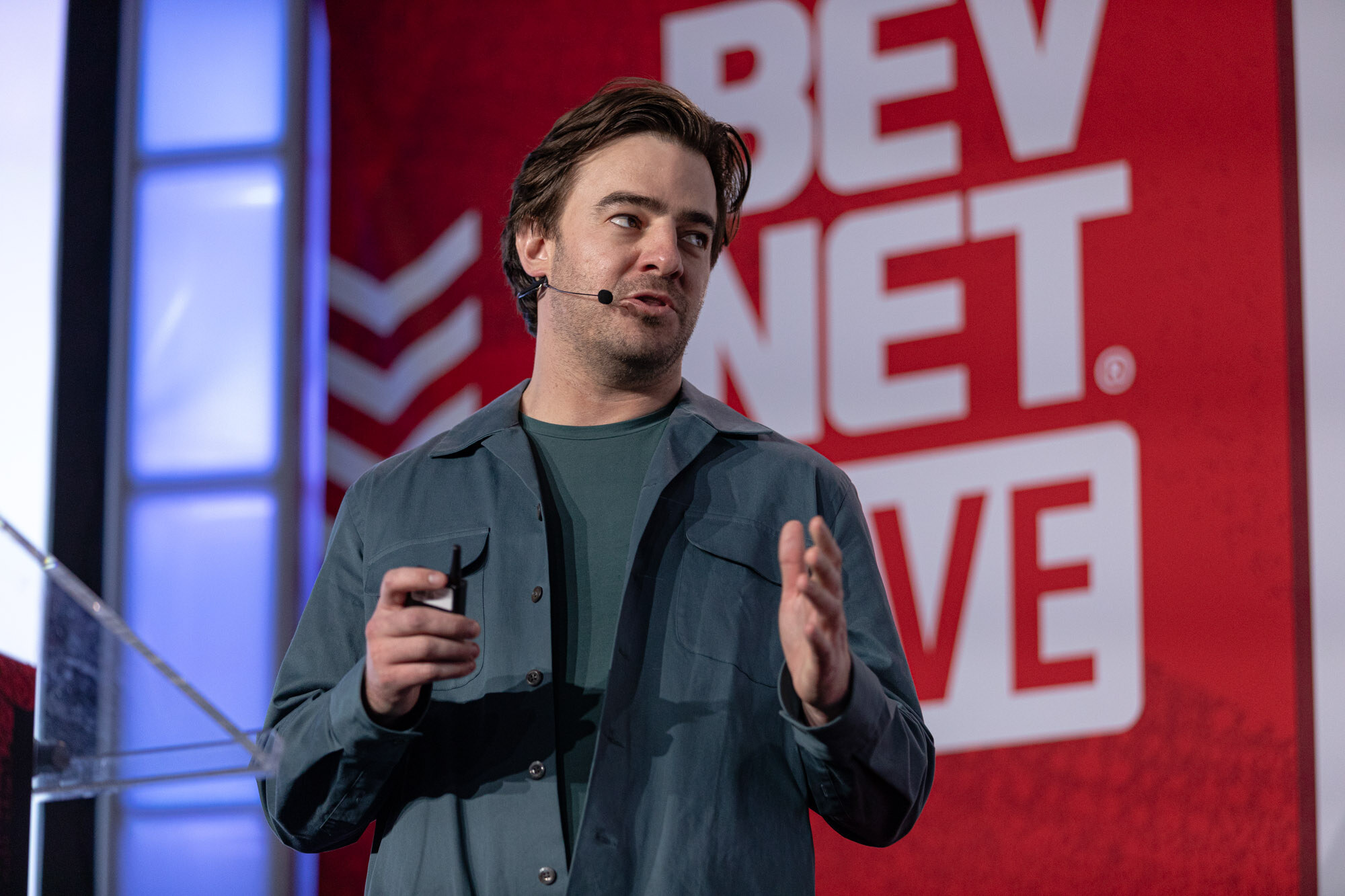 Man in a teal jacket gestures with both hands in front of a red BevNET Live backdrop.