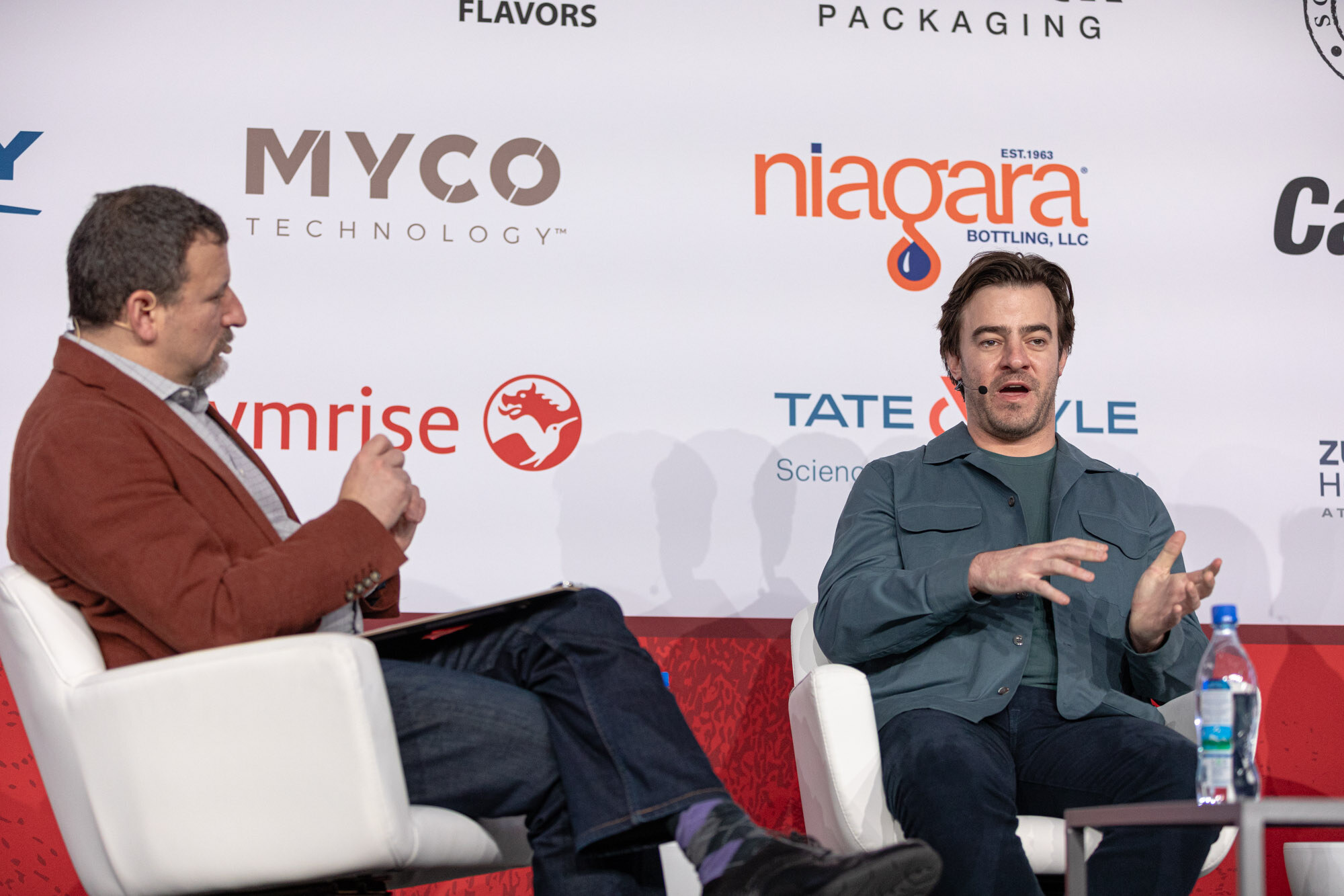 Two men sit in white armchairs on stage, one in a red jacket and one in a green jacket, in front of a sponsor wall.