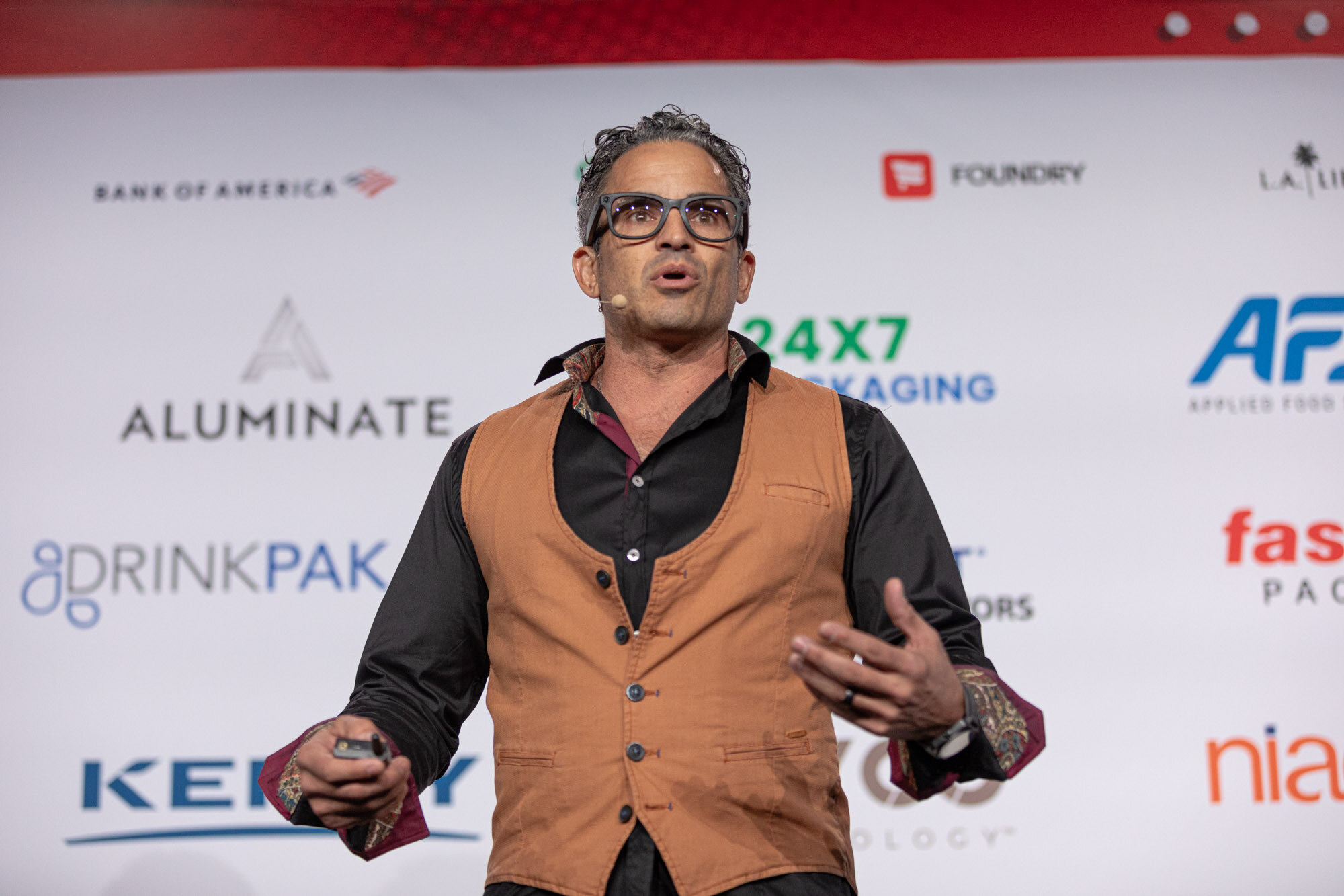 Man in an orange vest and glasses gestures with open hands on stage in front of a sponsor wall.