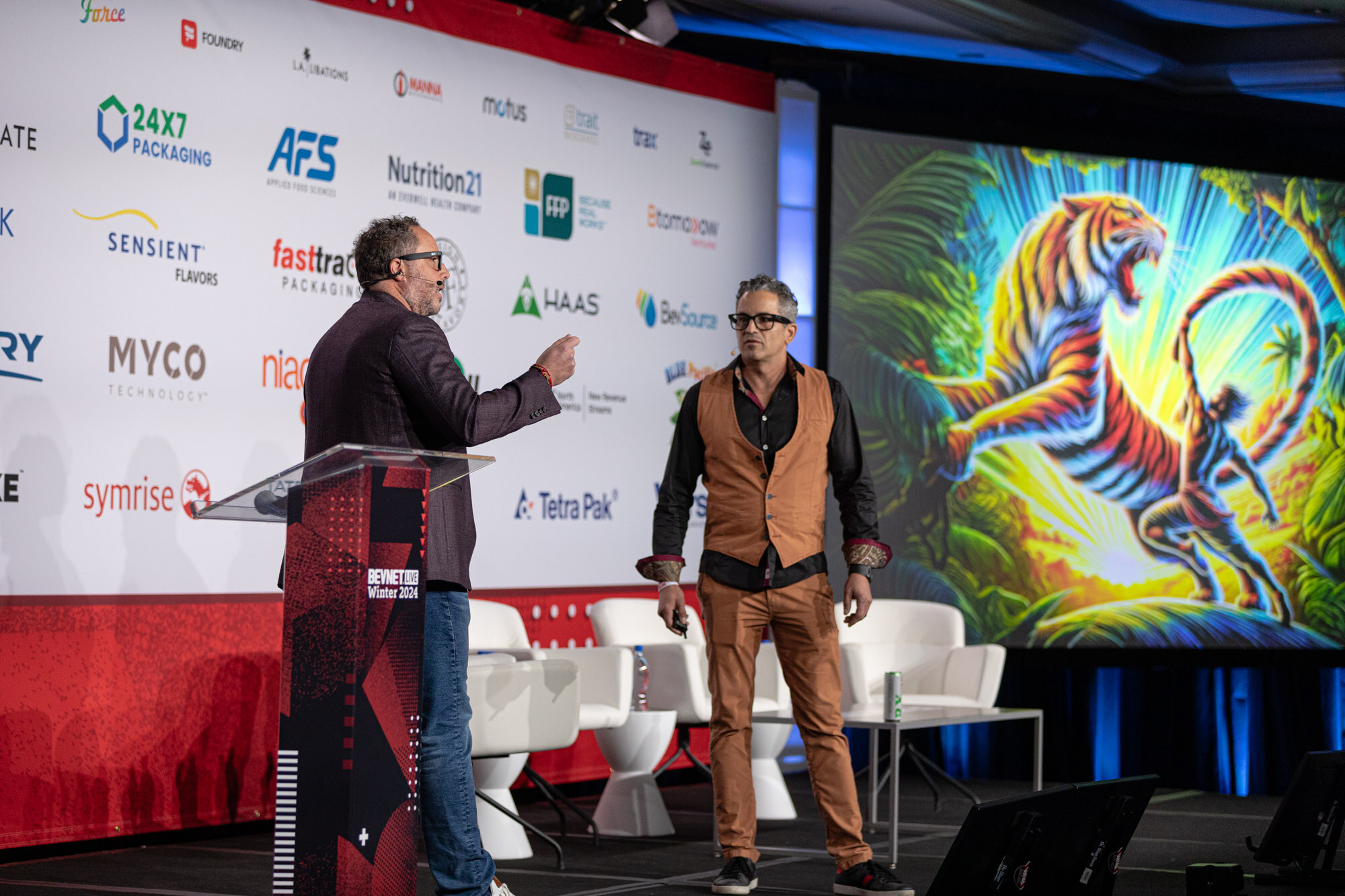 Two men stand on stage in front of a sponsor wall with a colorful tiger illustration on the screen.