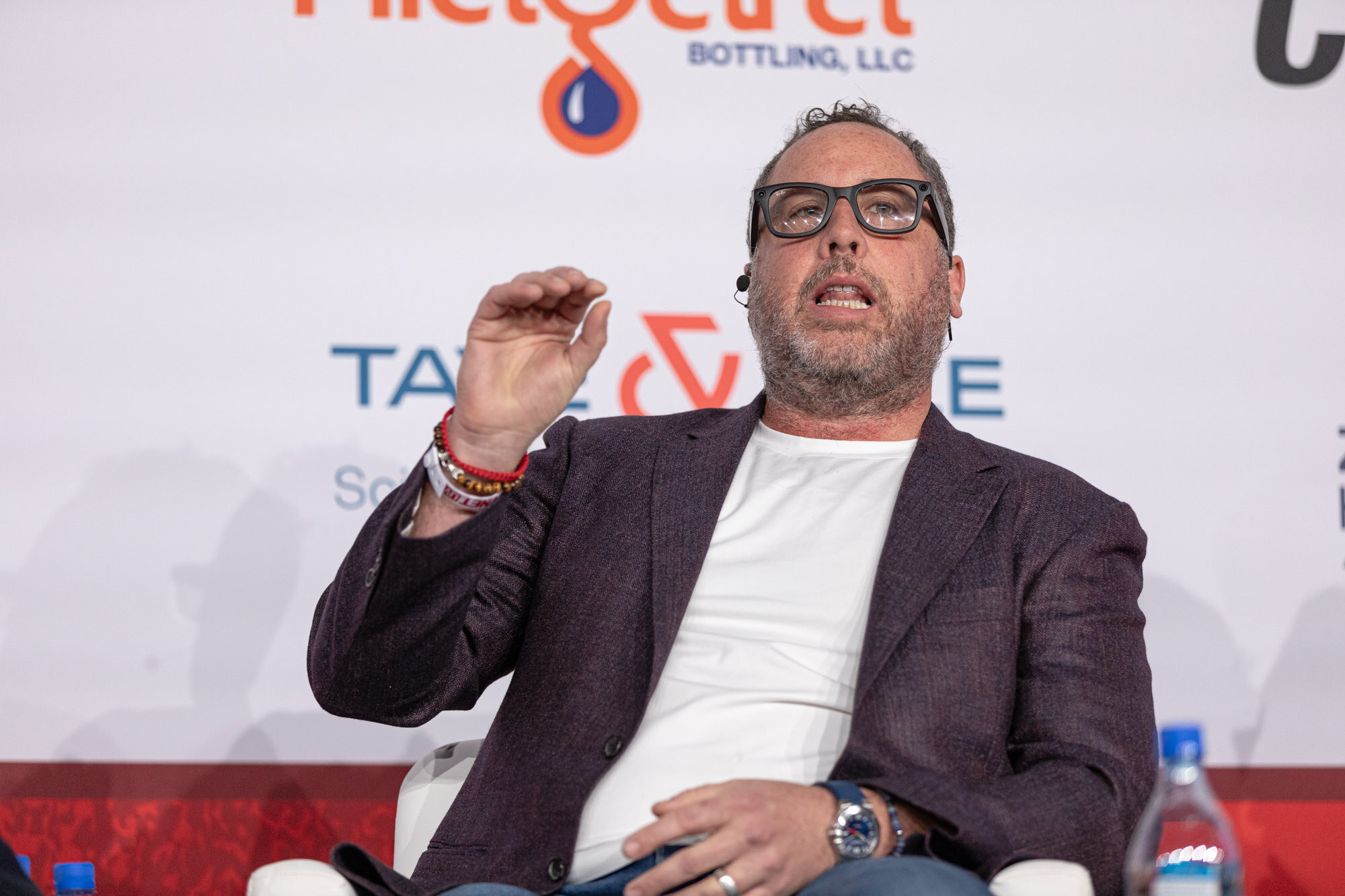 Bearded man with glasses in a dark blazer and white t-shirt raises one hand while seated in front of a sponsor wall.