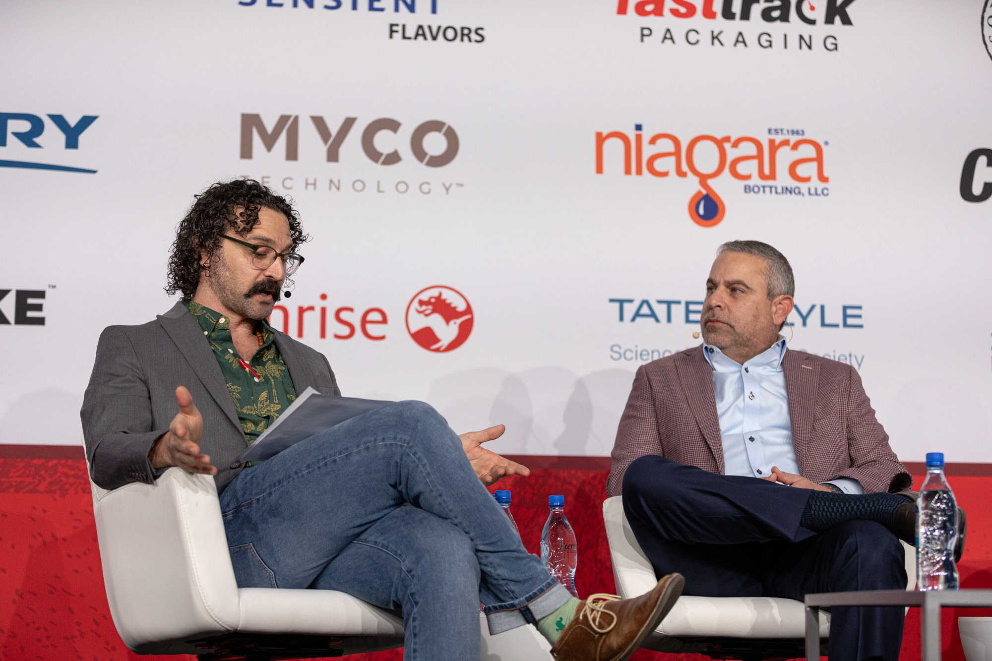 Two men sit in white armchairs on stage in front of a sponsor wall, one with curly hair and glasses and one in a plaid blazer.