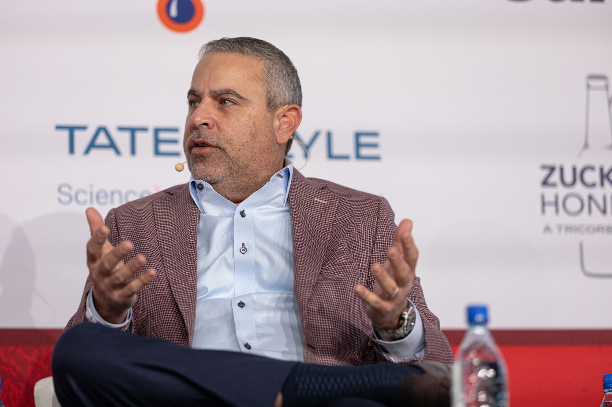 Man in a red checked blazer and light blue shirt gestures with both hands while seated on stage.