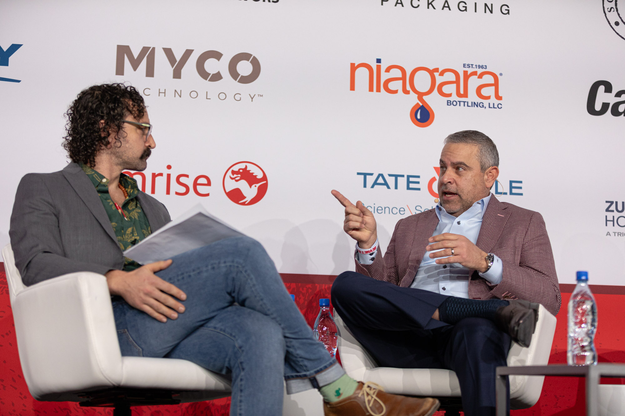 Two men seated in white armchairs, one pointing toward the other, in front of a sponsor wall.