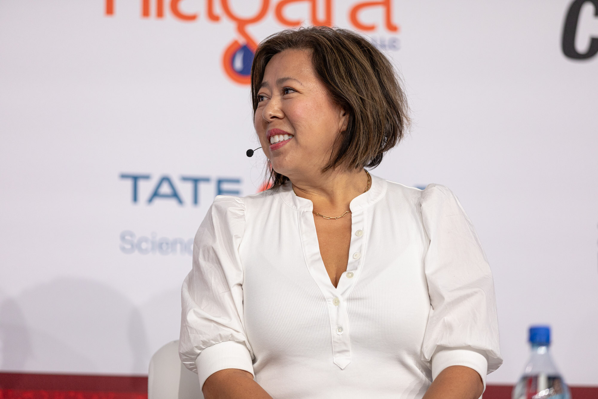 Smiling woman in a white blouse sits in front of a sponsor wall during an interview.
