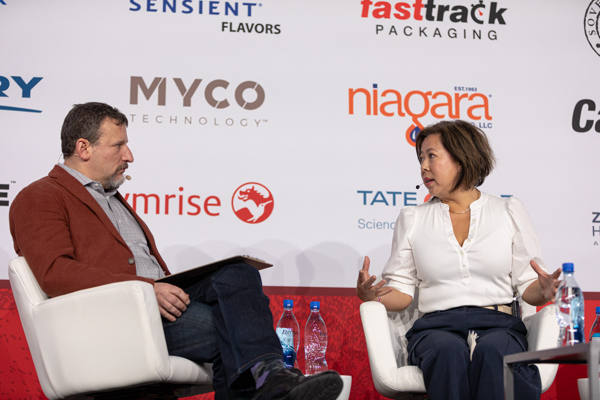 A man in a red blazer and a woman in a white blouse sit in white armchairs on stage.