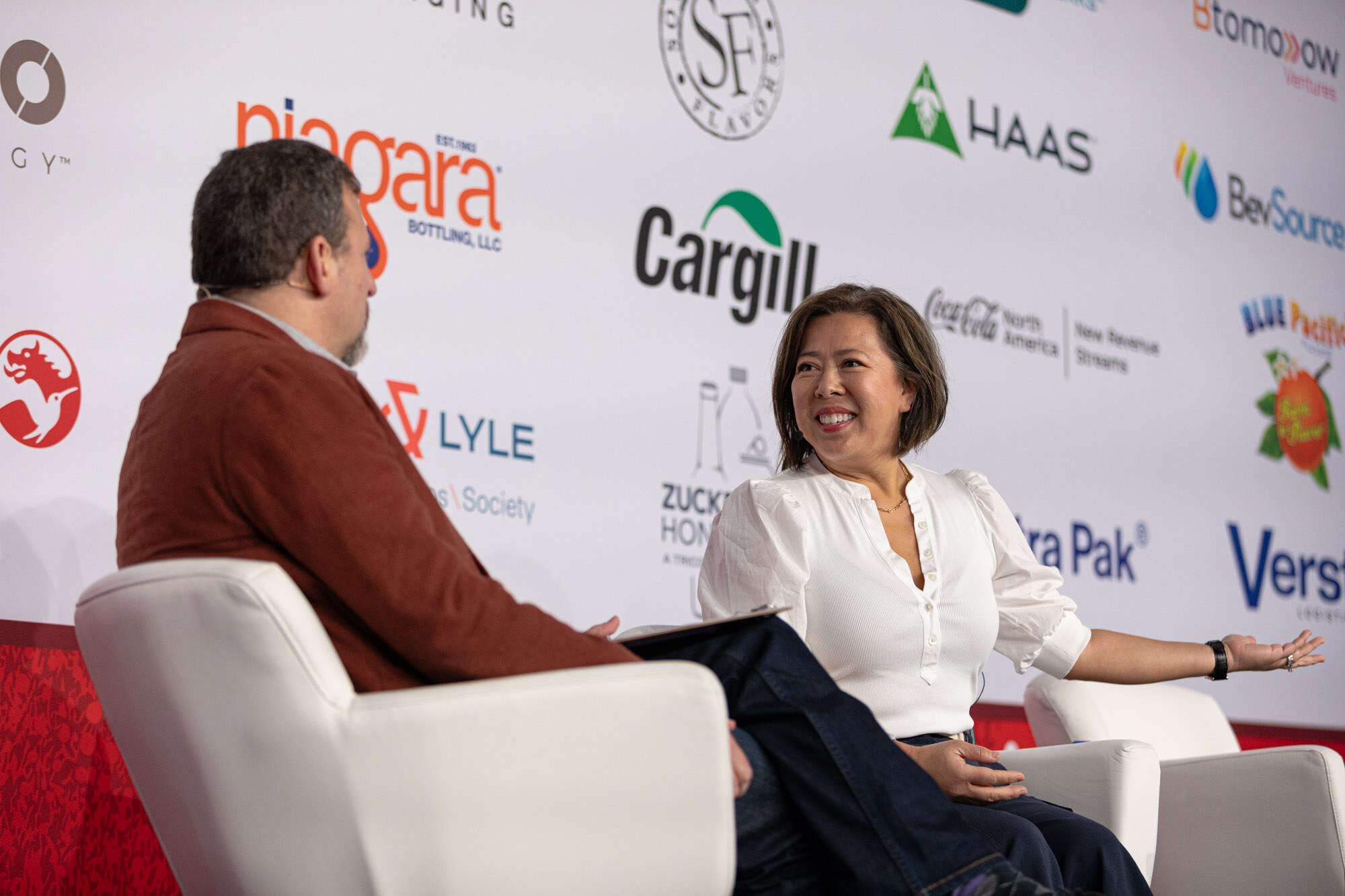 Woman in white smiles broadly while seated across from a man in a red blazer in front of a sponsor wall.