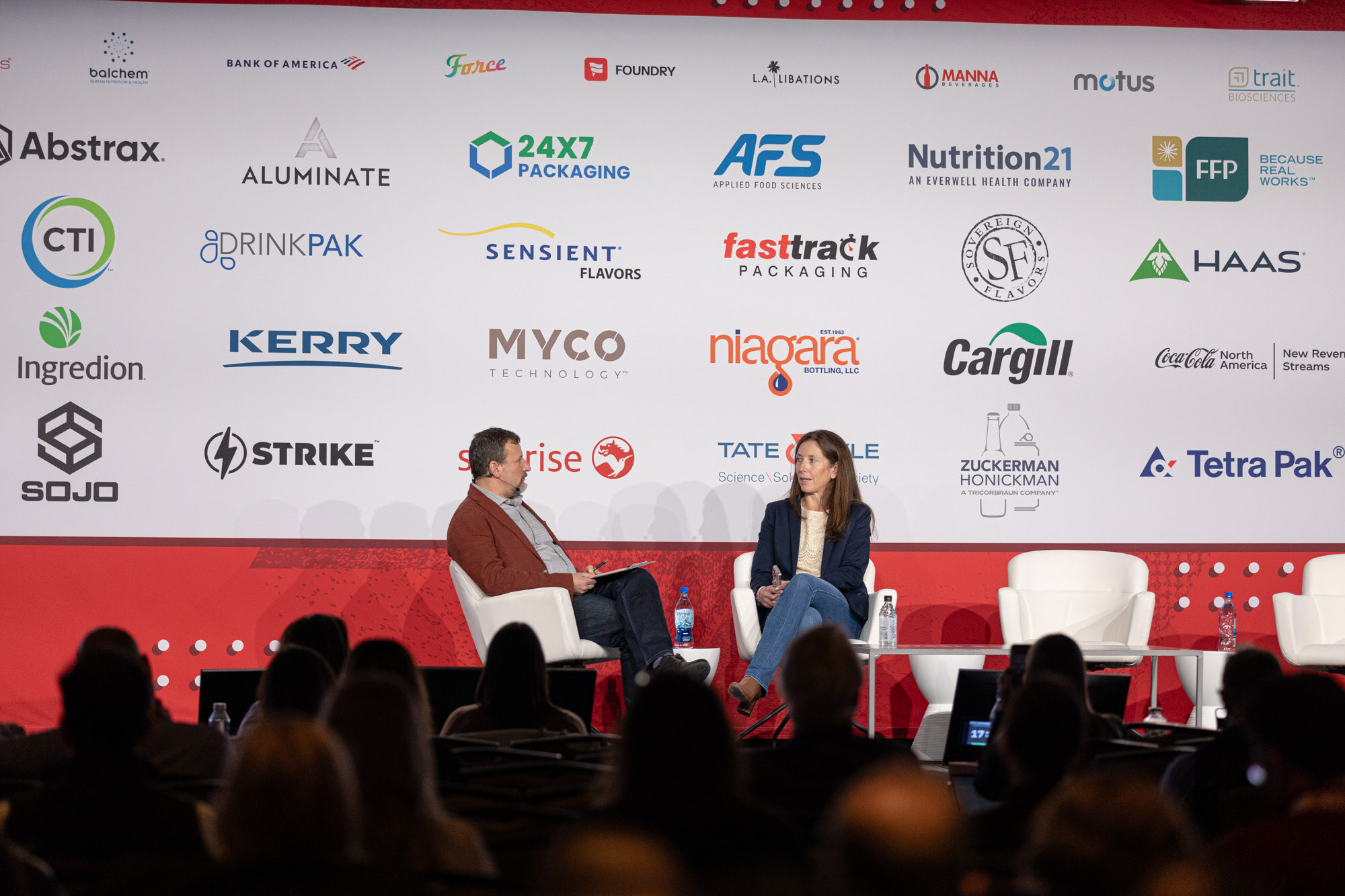 Two people sit in white chairs on a stage in front of a sponsor wall with audience heads in the foreground.
