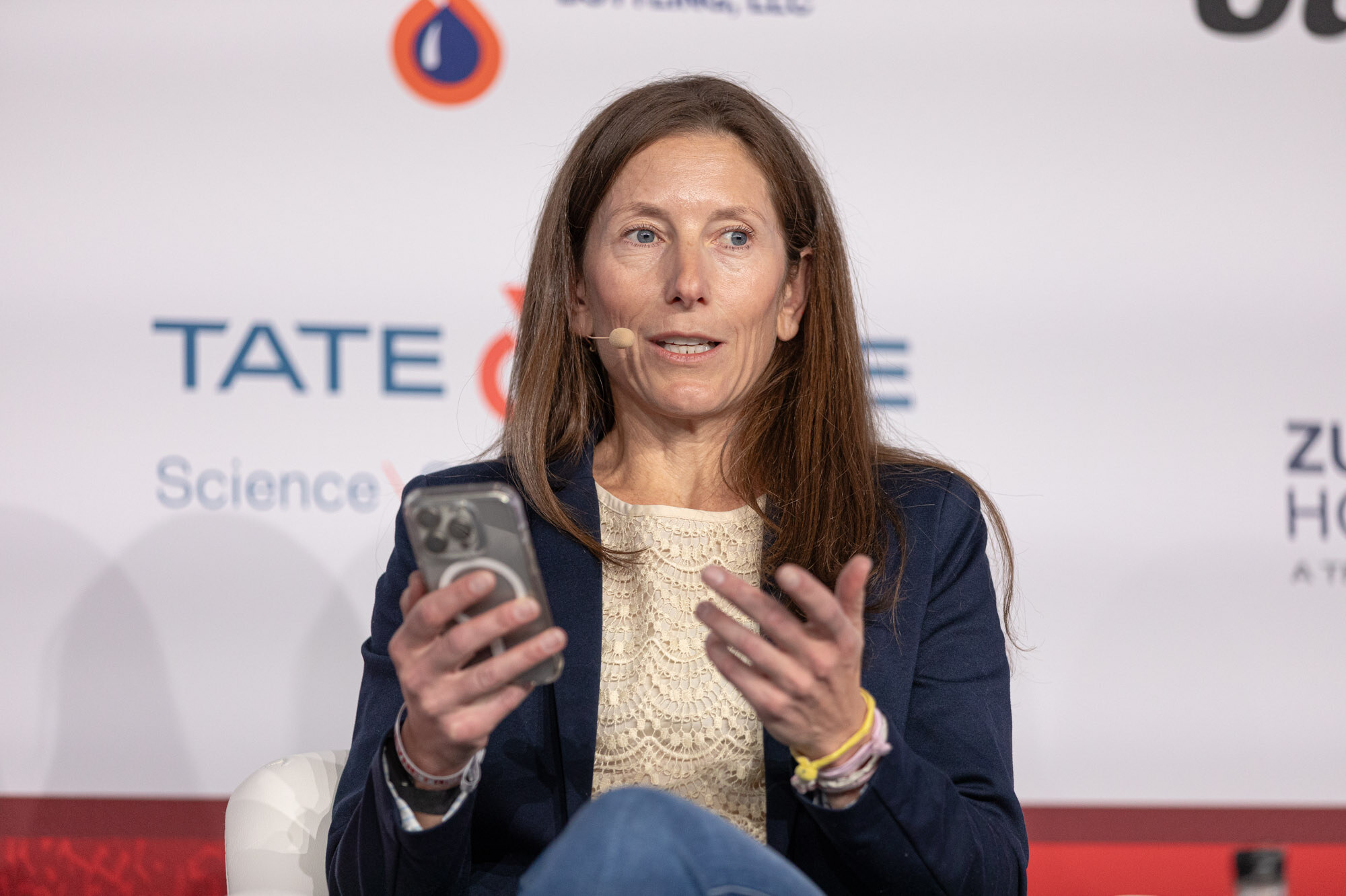 Woman with long brown hair in a navy blazer holds a phone and talks while seated in front of a sponsor wall.